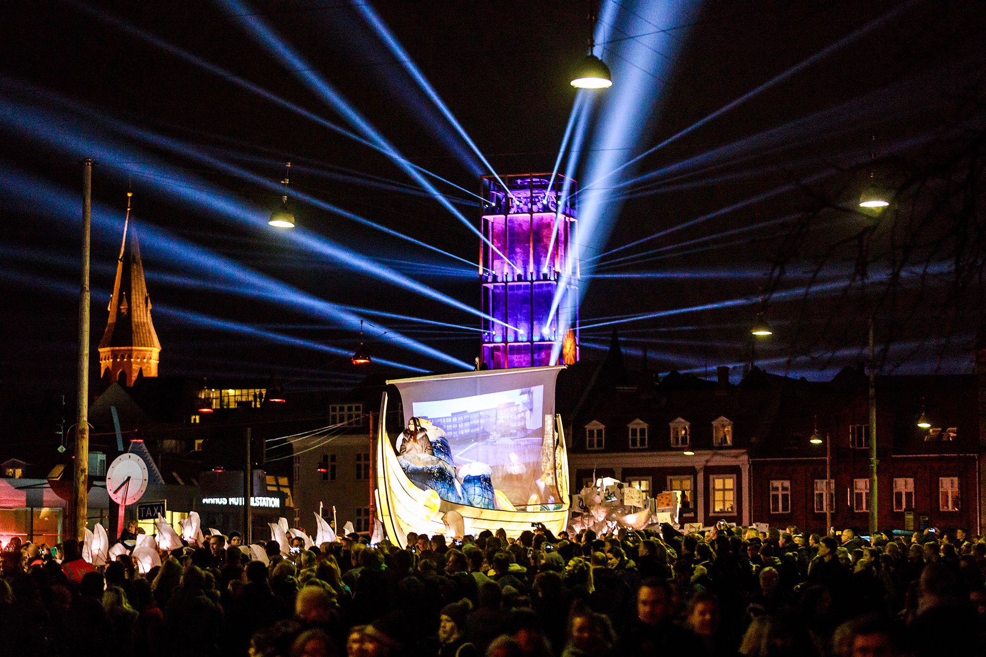A city square at night filled with people, an illuminated tower structure and a lit boat-shaped lantern in front of a church spire.