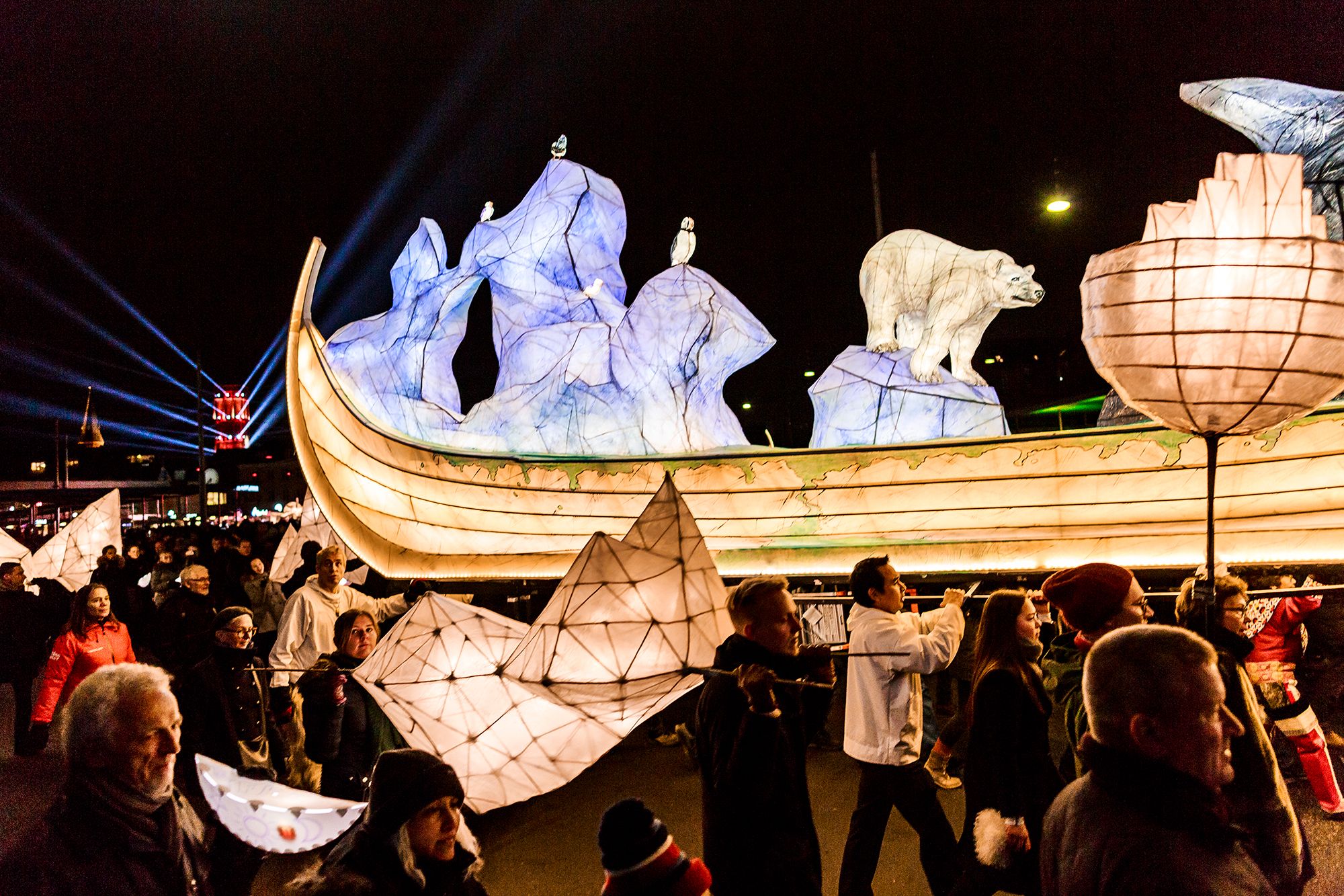 A large illuminated lantern in the shape of a wooden ship carrying pale animal figures, carried through a night-time crowd.