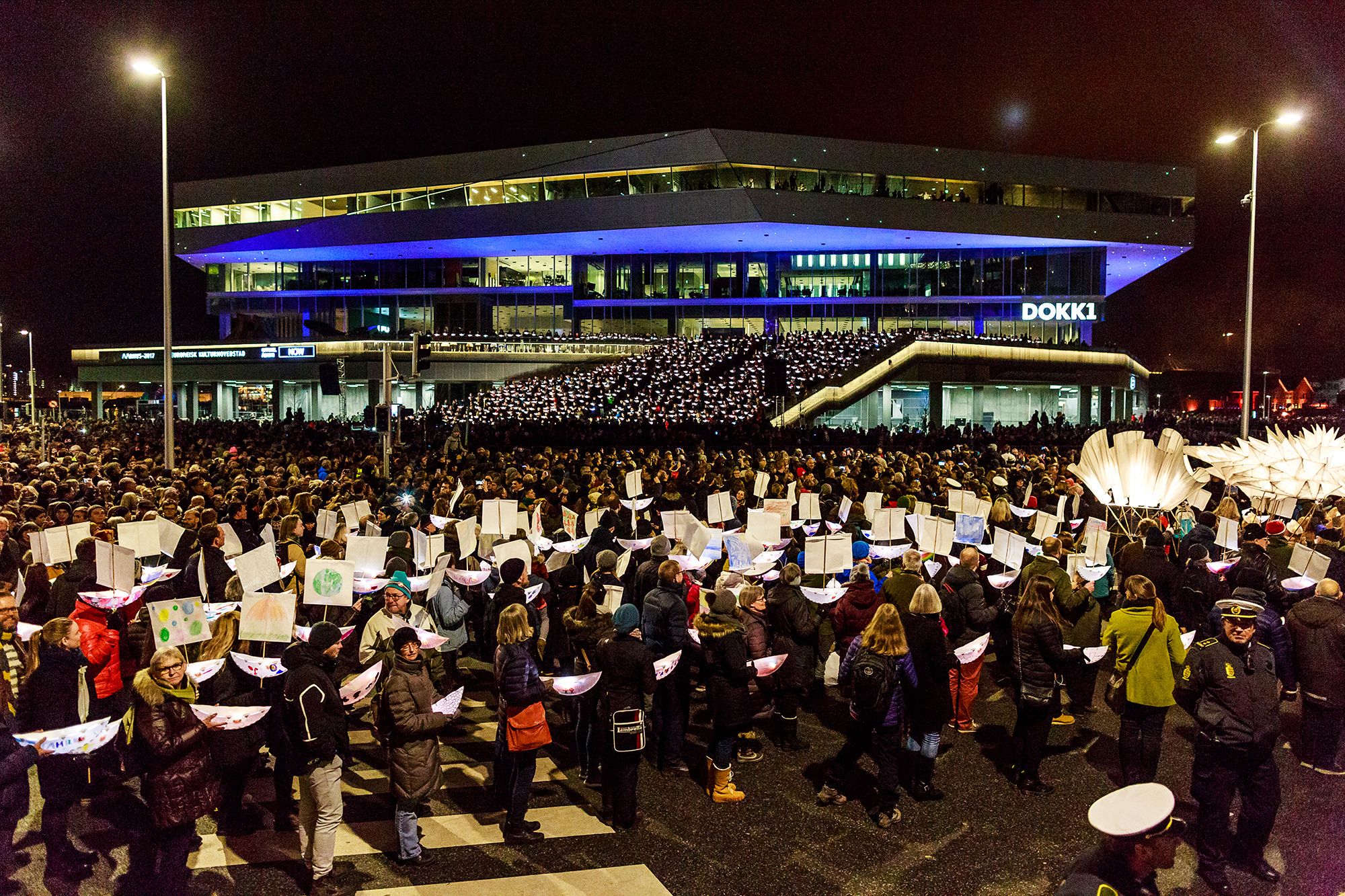 A dense night-time crowd in front of a large illuminated modern building, many people holding tall white paper lanterns on poles.