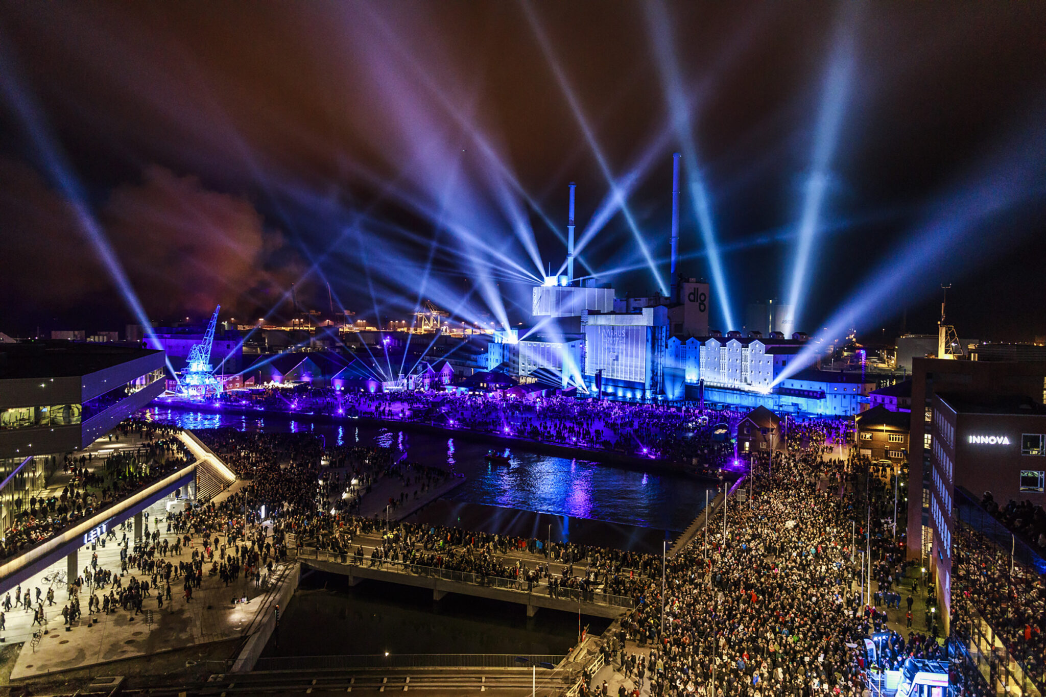 Aerial view of a harbour at night, blue and white beams crossing above a floodlit stage with a dense crowd lining both quaysides.