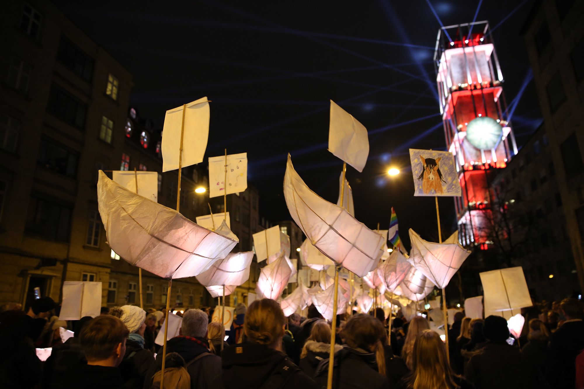 Close view of tall white paper lanterns shaped like sails, held up on poles against a dark sky beside a red and white tower.