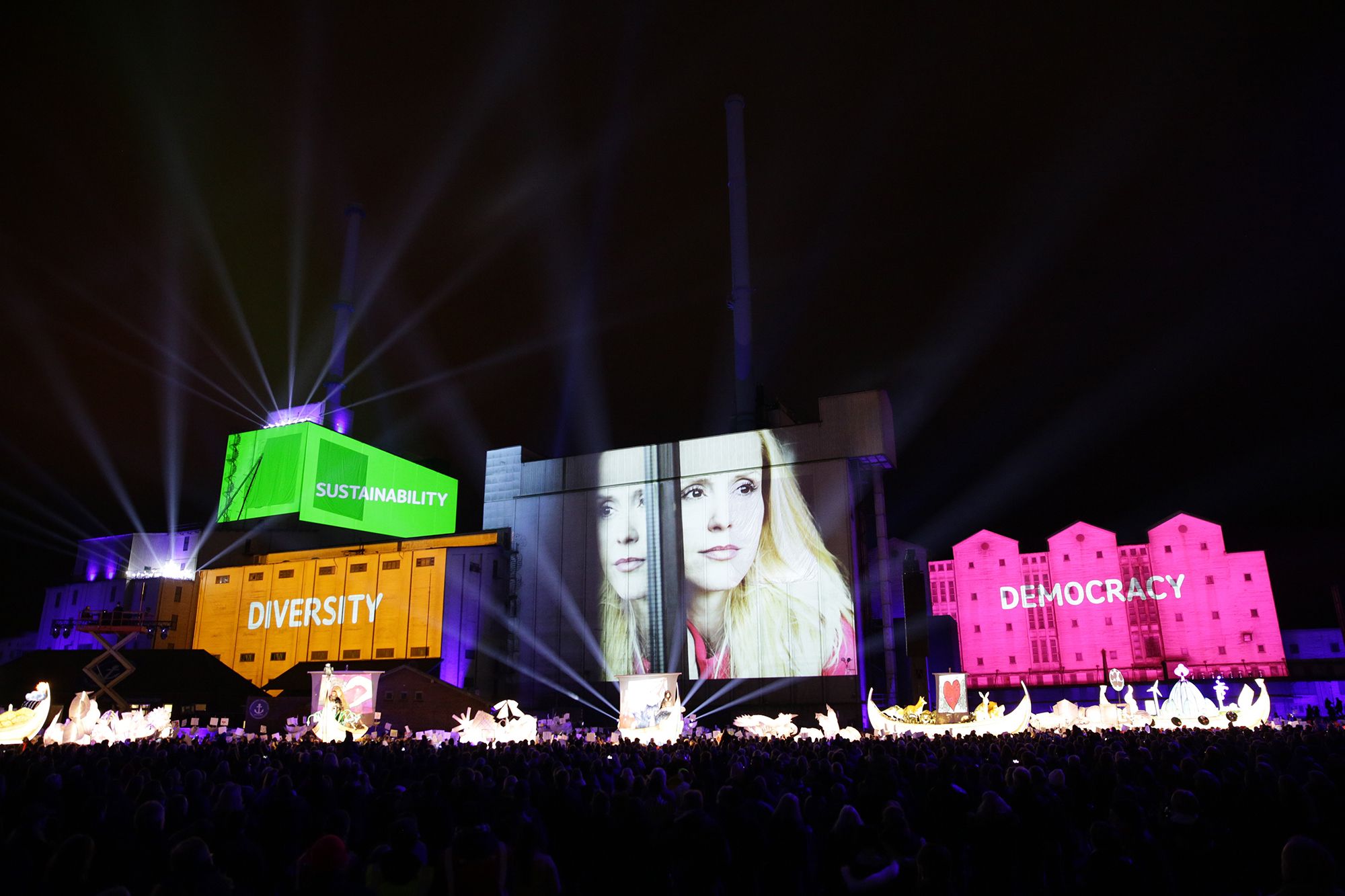A long outdoor stage at night carrying projected images of faces, a green container and brightly coloured panels of text.