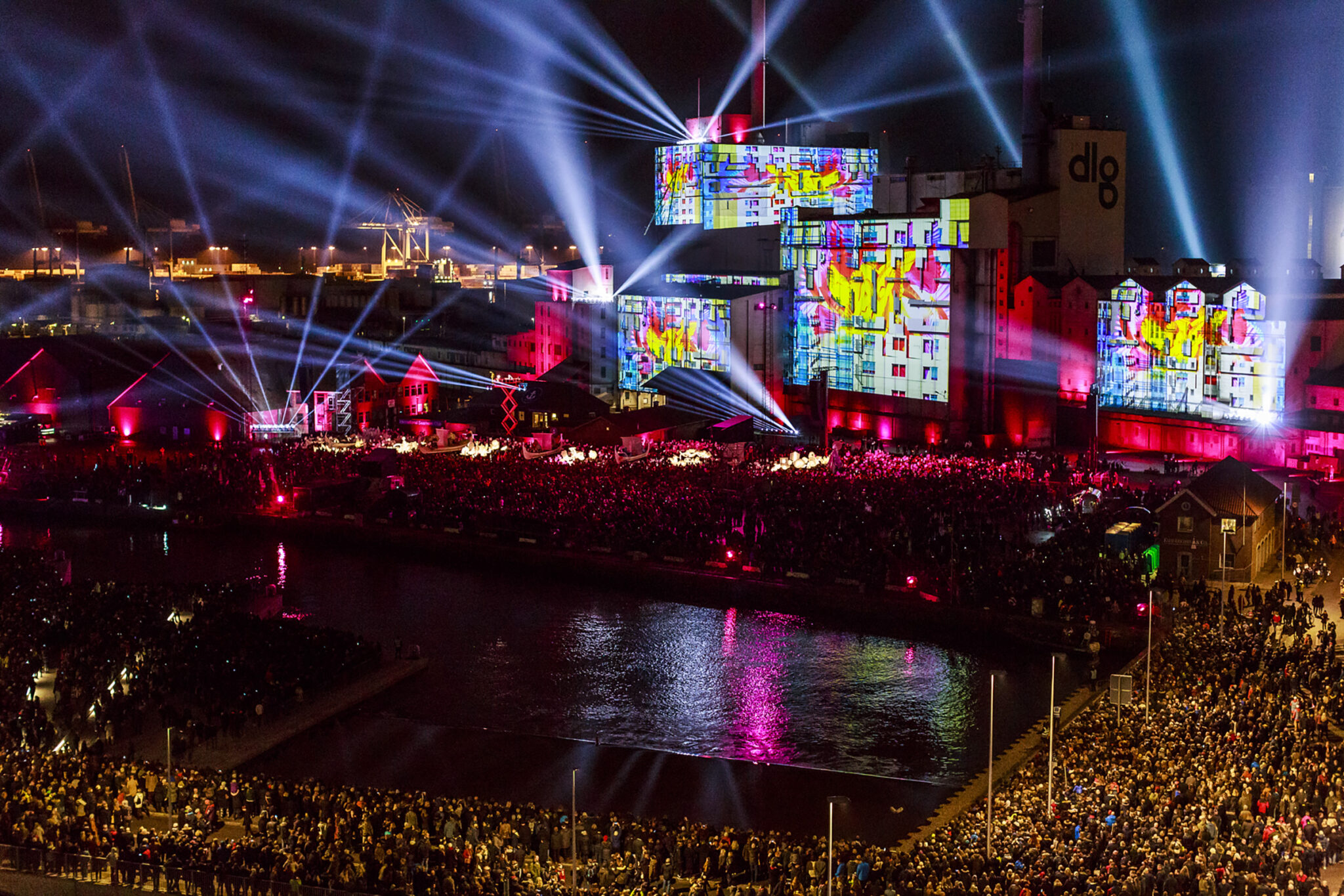 Aerial night view of a harbour, a long building facade covered in brightly coloured projections, pink and red light reflecting on the water.