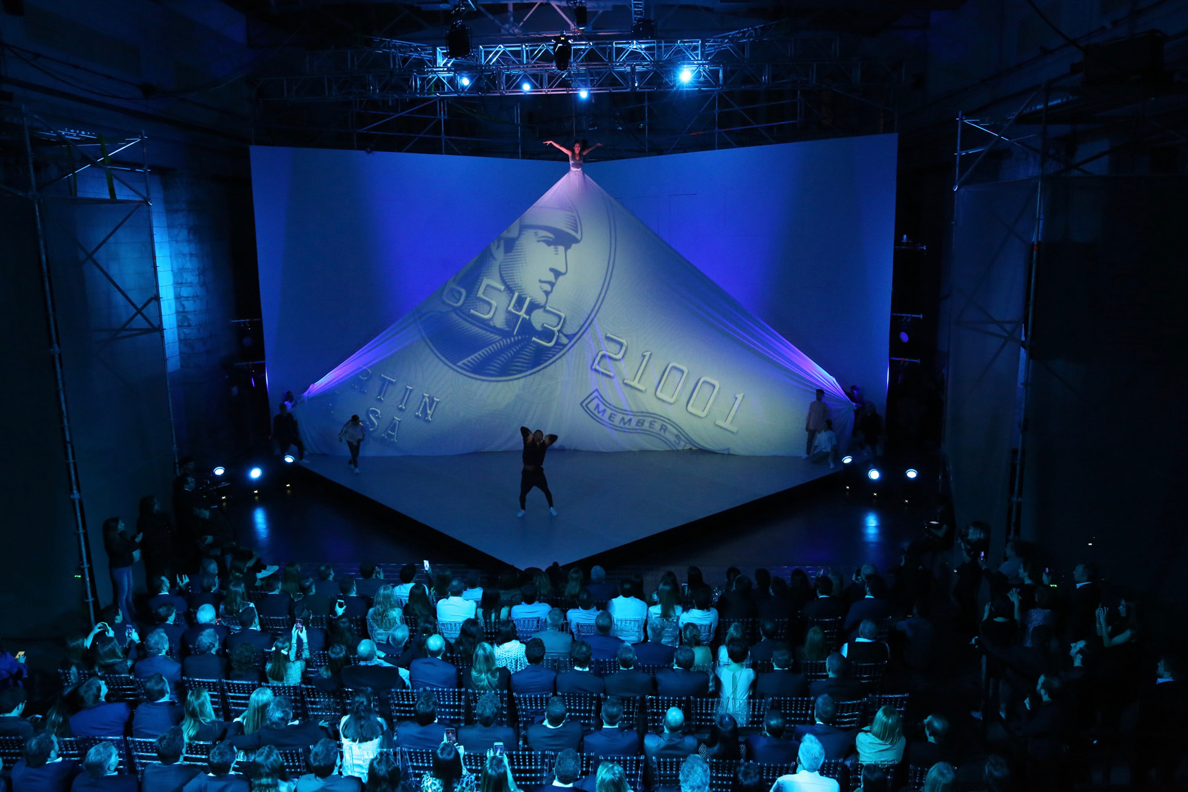 A seated audience in a dark venue watches a large angled projection screen lit in blue, with a performer standing on the stage below.