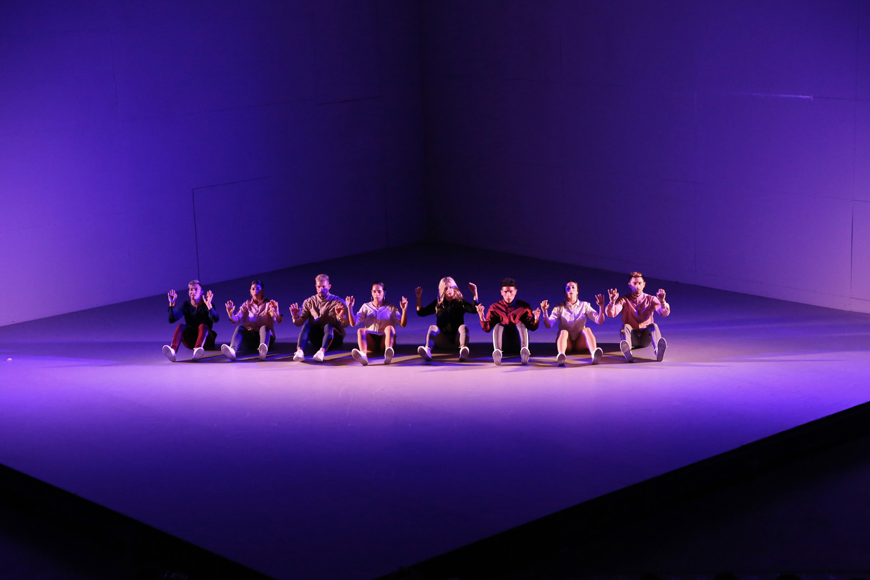 A line of dancers sits in a row at the back of an empty stage washed in purple light.