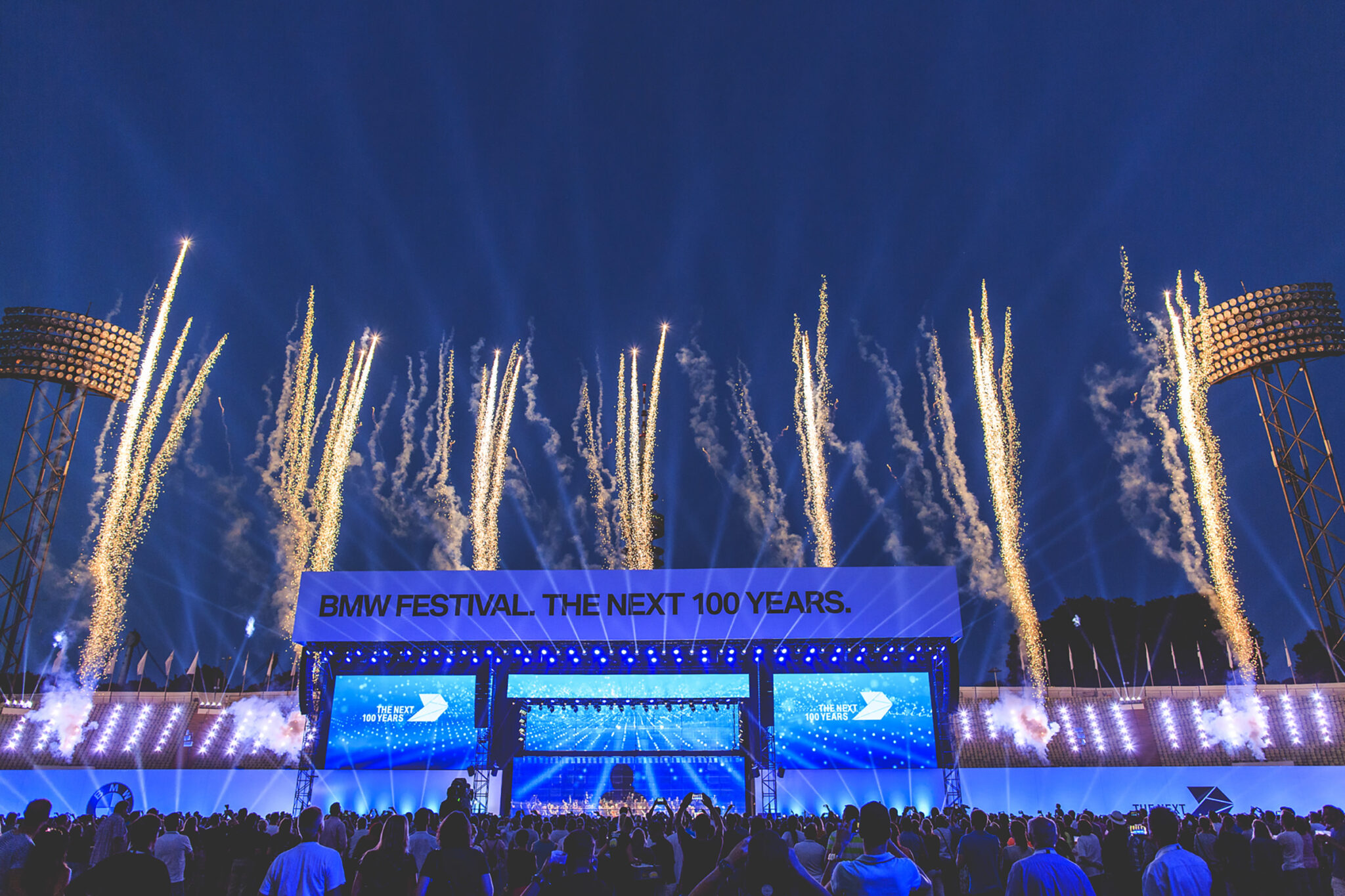 Rows of golden fireworks rise behind a wide stage lit in blue at dusk, with three large screens and a crowd watching from the field.