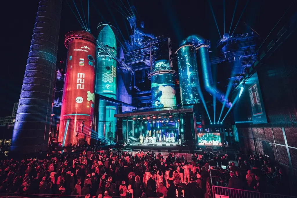 A large crowd gathered at an industrial venue with illuminated silos and colorful light projections on towering structures, creating a vibrant atmosphere. Battle Royal Studios