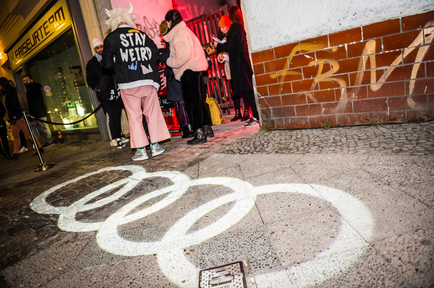 A group of people gathers outside a venue at night, with one person wearing a jacket that says "Stay Weird." The ground is illuminated with the projection of four interlocking rings. Battle Royal Studios