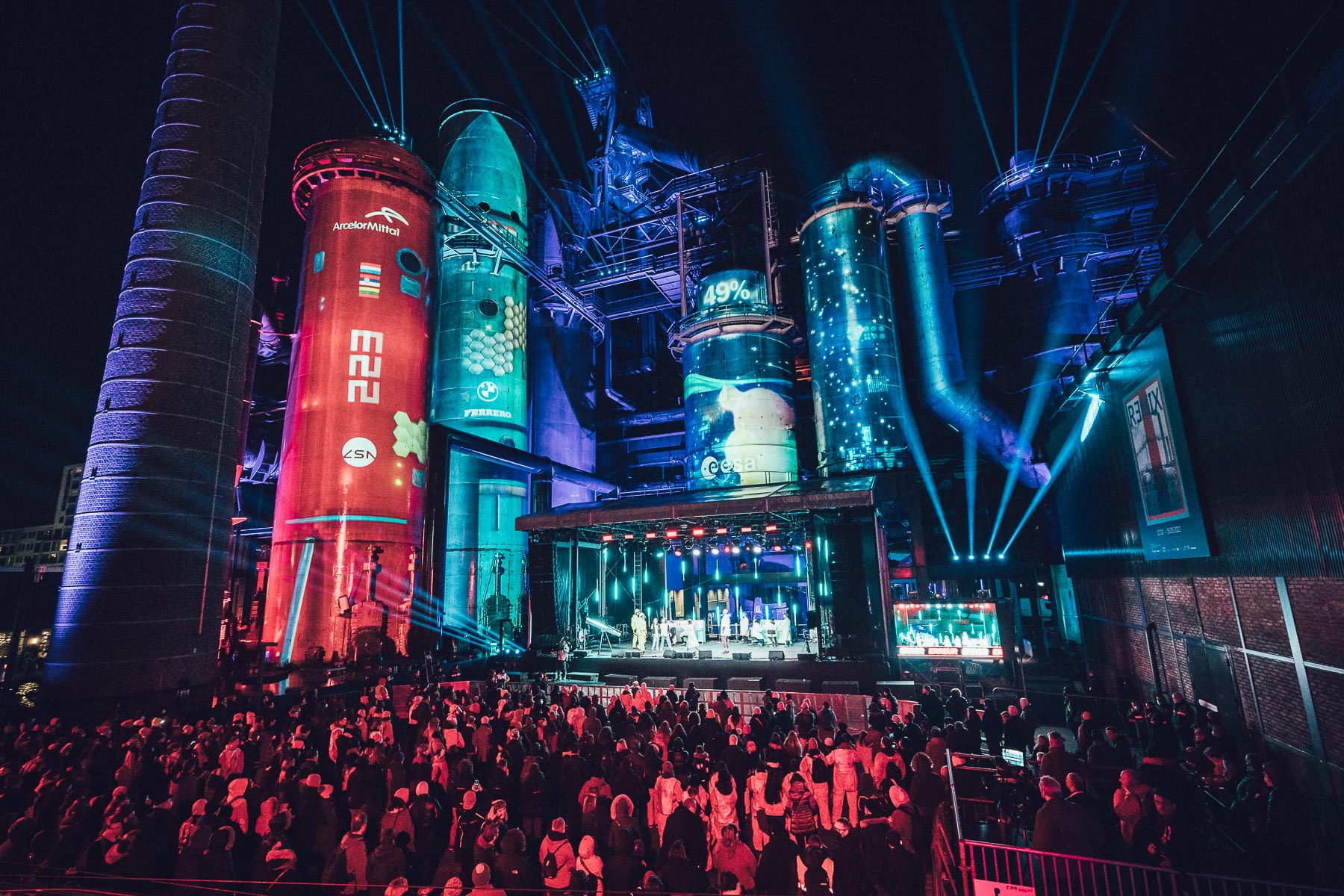 A tall industrial structure at night with its towers and silos lit in red, teal and blue projected imagery, with beams of light overhead and a large crowd gathered in front of a lit stage at its base.