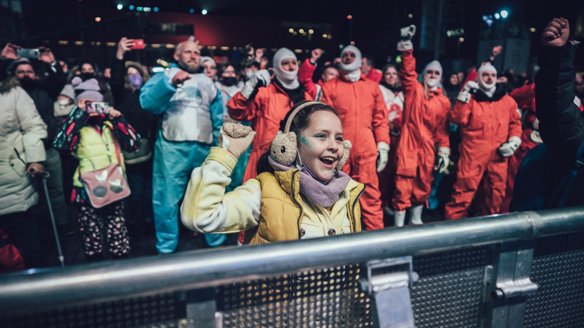 A child in ear defenders laughs and raises a fist behind a crowd barrier at night, surrounded by people in red and orange boiler suits.