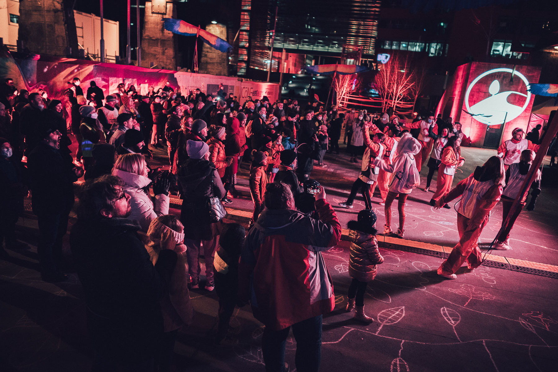 A large crowd stands outdoors at night on a square lit in deep red and pink, watching performers in orange boiler suits dance on chalk-drawn paving, with painted banners and a lit building behind.