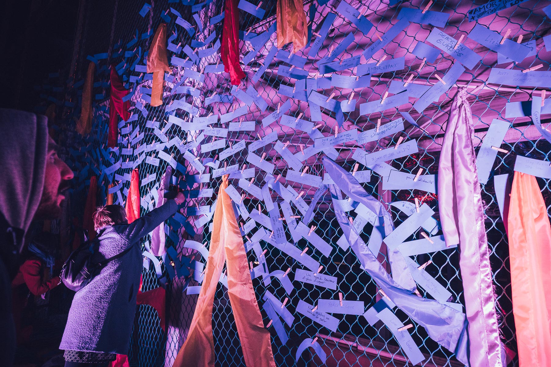 A wire mesh fence densely covered in handwritten paper notes held by clothes pegs, interspersed with hanging fabric ribbons, lit in pink and violet, with two people pinning a note at one side.