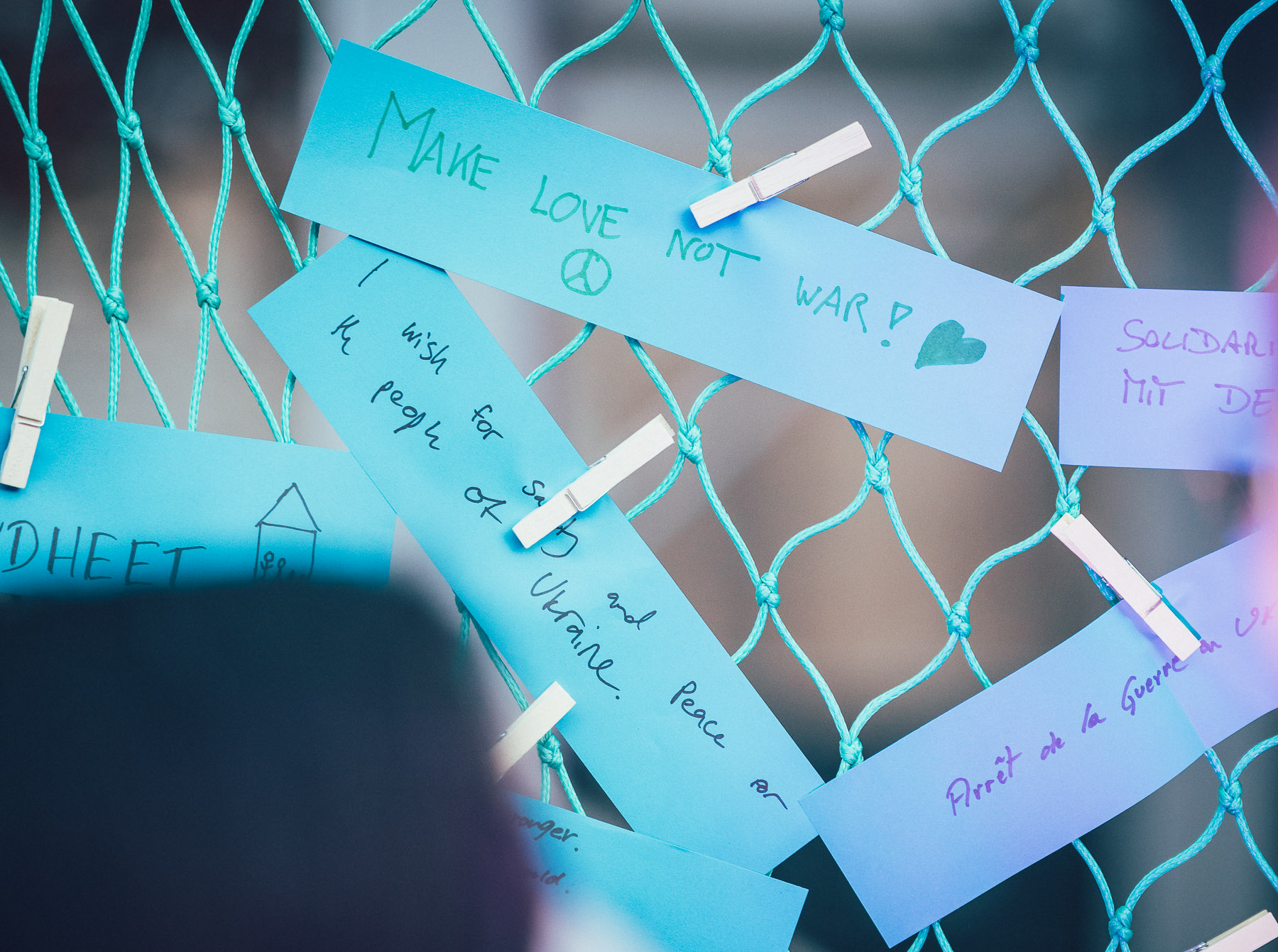 Pale turquoise cards covered in handwriting hang from a wire mesh fence, lit from one side so the writing reads against the dark background.