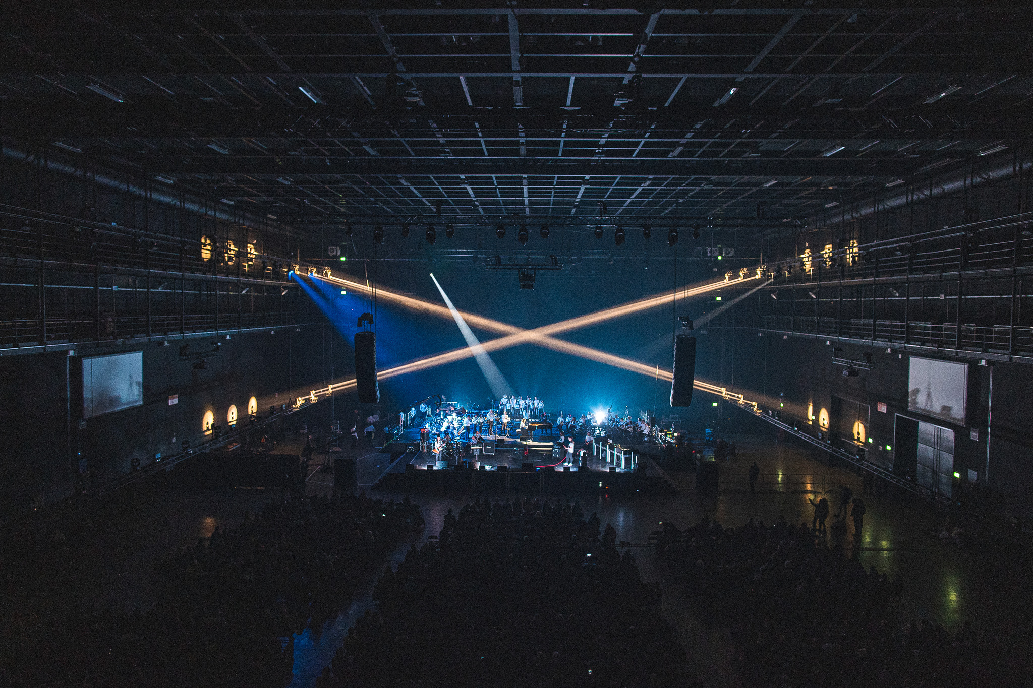 Two wide beams of warm white light cross in an X above a large dark arena, with a blue beam behind them and musicians lit on a stage in front of a seated audience.