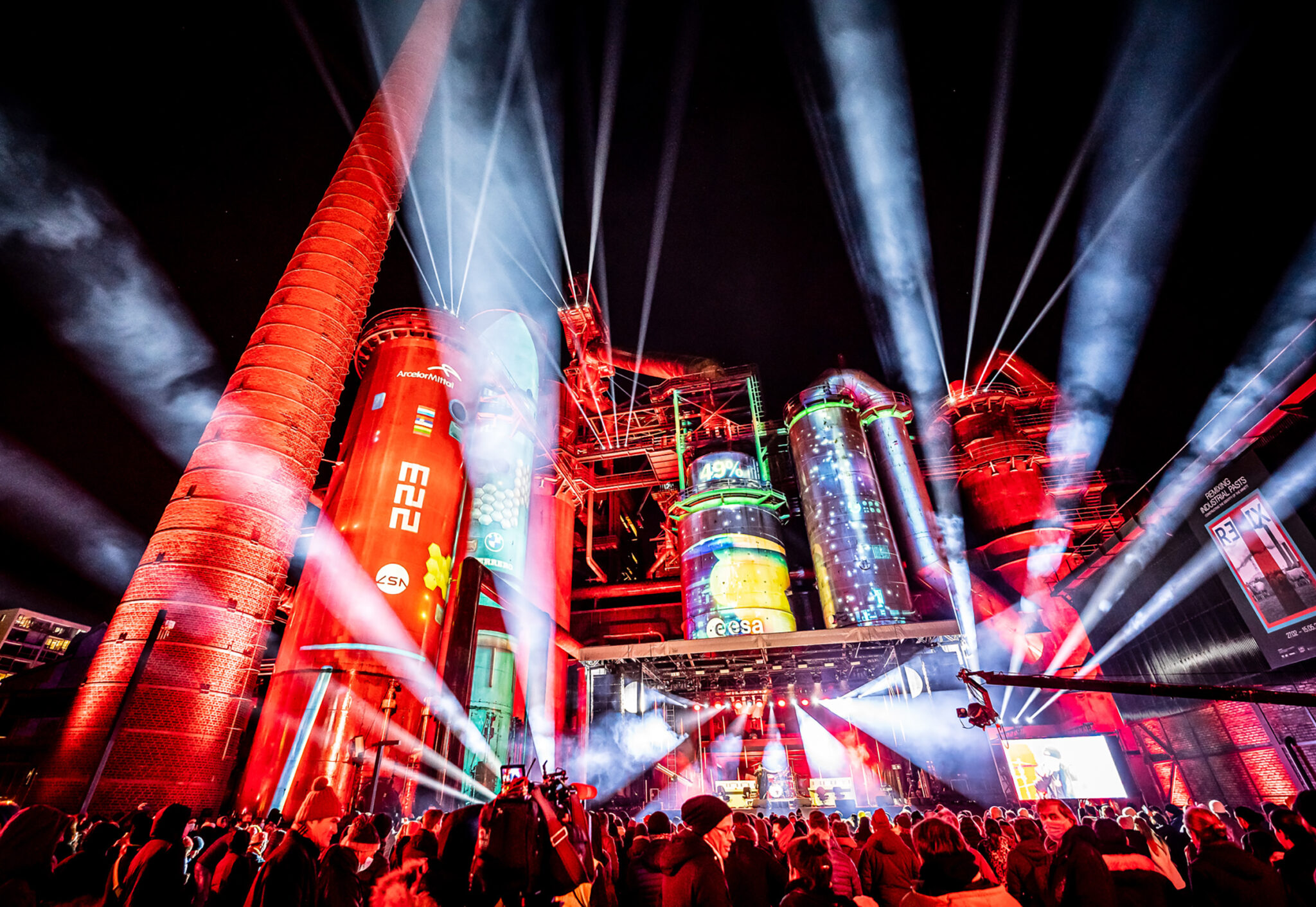 Red and white beams of light fan out above an industrial structure at night, with a crowd silhouetted in front of a lit stage area.