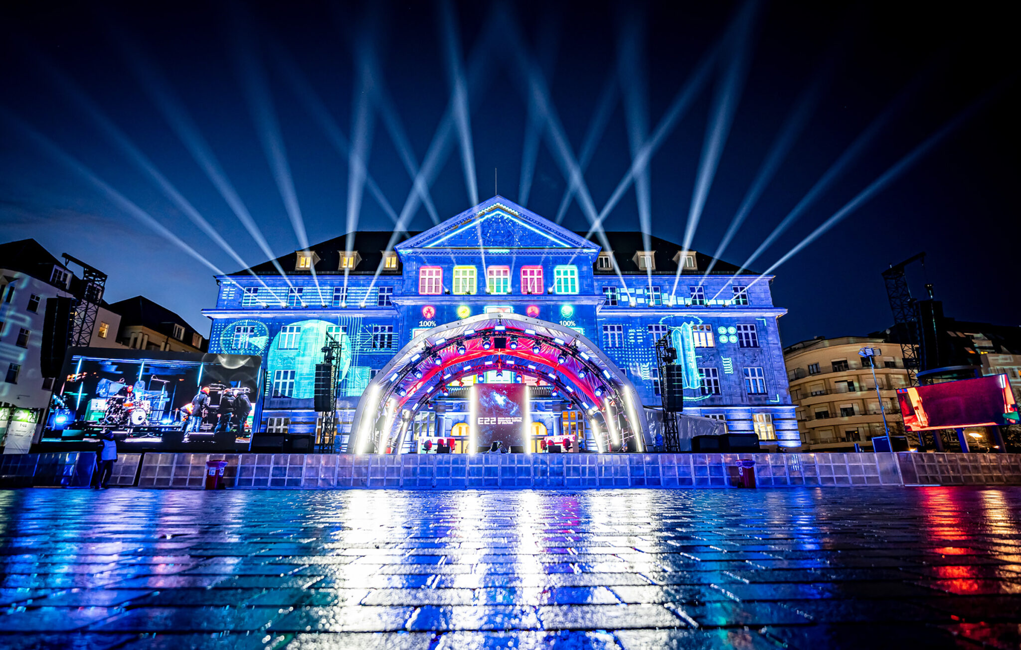 A large neoclassical building at night with its full facade covered in blue and white projected patterns, mirrored in still water across the square in front of it.