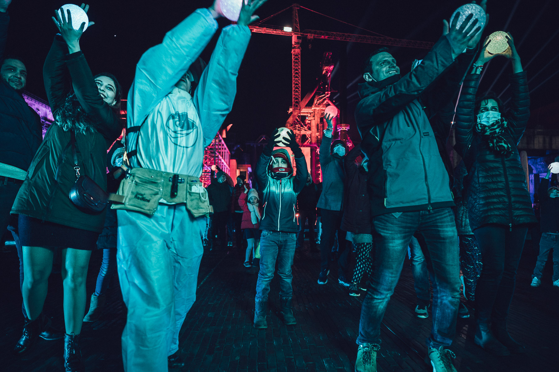 A crowd outdoors at night reaches upward holding glowing white spheres above their heads, lit in turquoise, with a red-lit crane and industrial structure behind them.