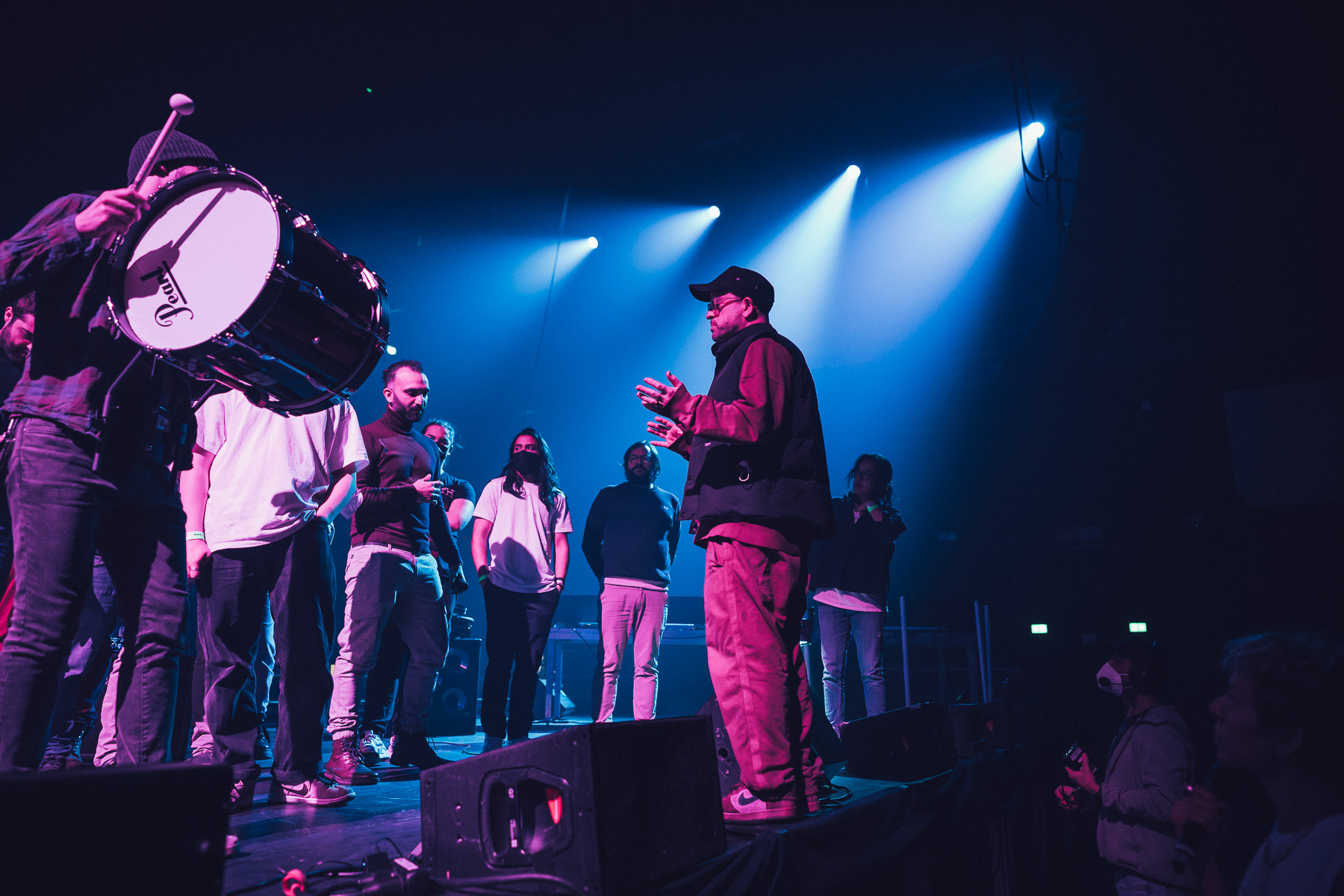 A group of musicians and crew stand on a stage lit in blue and magenta, one holding a large bass drum with a mallet raised, while a person in a cap gestures as they speak to them.
