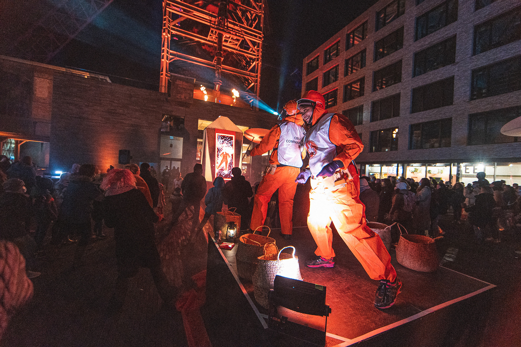 Two performers in orange boiler suits, reflective tabards and red helmets dance on a small low stage at night, surrounded by a standing crowd, with an industrial tower lit in orange behind them.