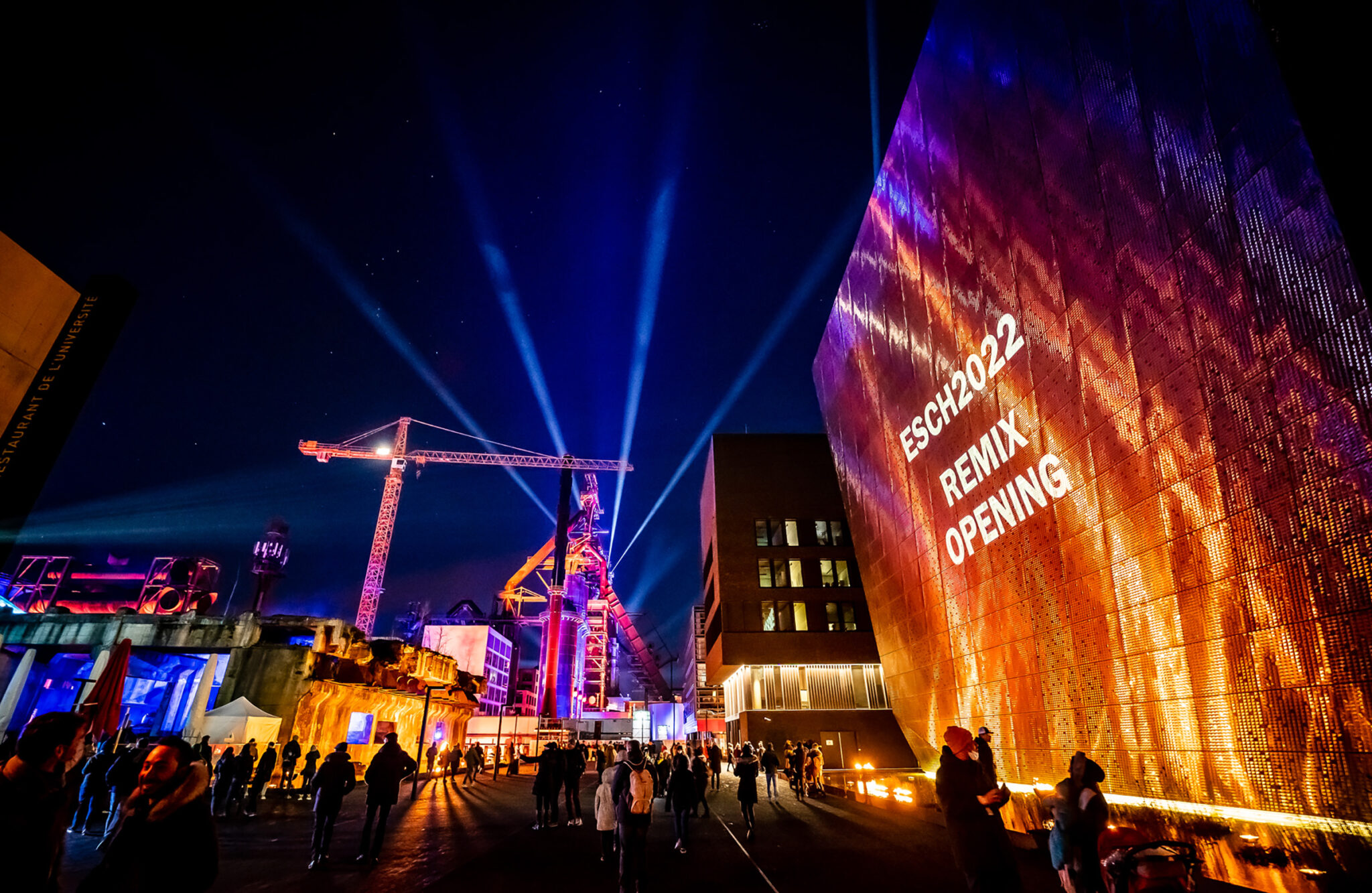 A tall building facade lit in deep orange with a crane silhouetted beside it, blue beams crossing the sky and a crowd filling the street below.