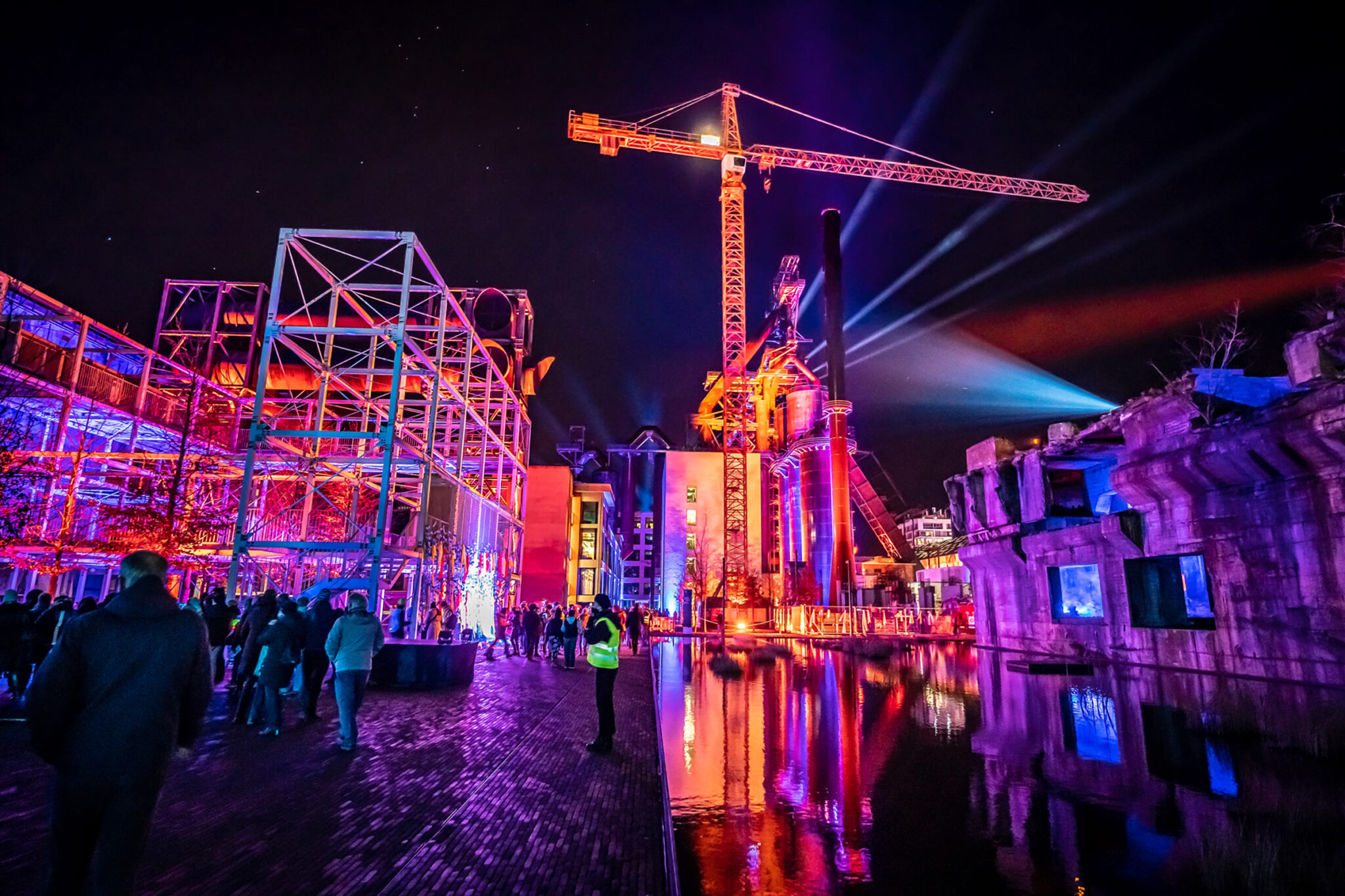 A tall industrial structure at night with its towers and silos lit in red, teal and blue projected imagery, with beams of light overhead and a large crowd gathered in front of a lit stage at its base.