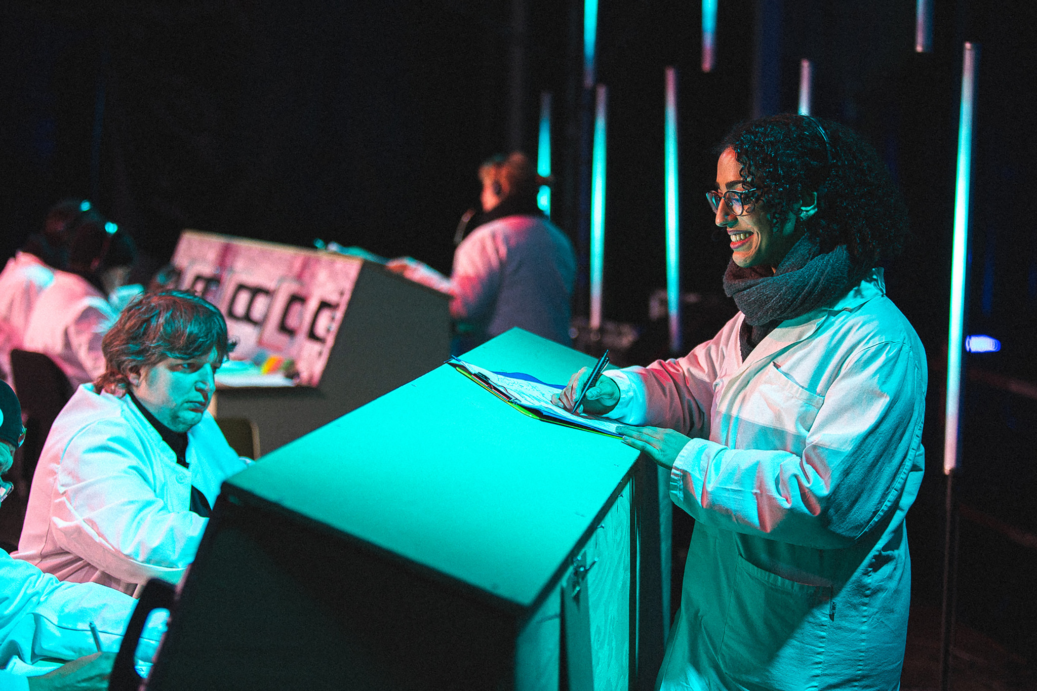 People in white lab coats stand at glowing turquoise desks writing on clipboards, with vertical light tubes and a printed panel behind them in a darkened space.