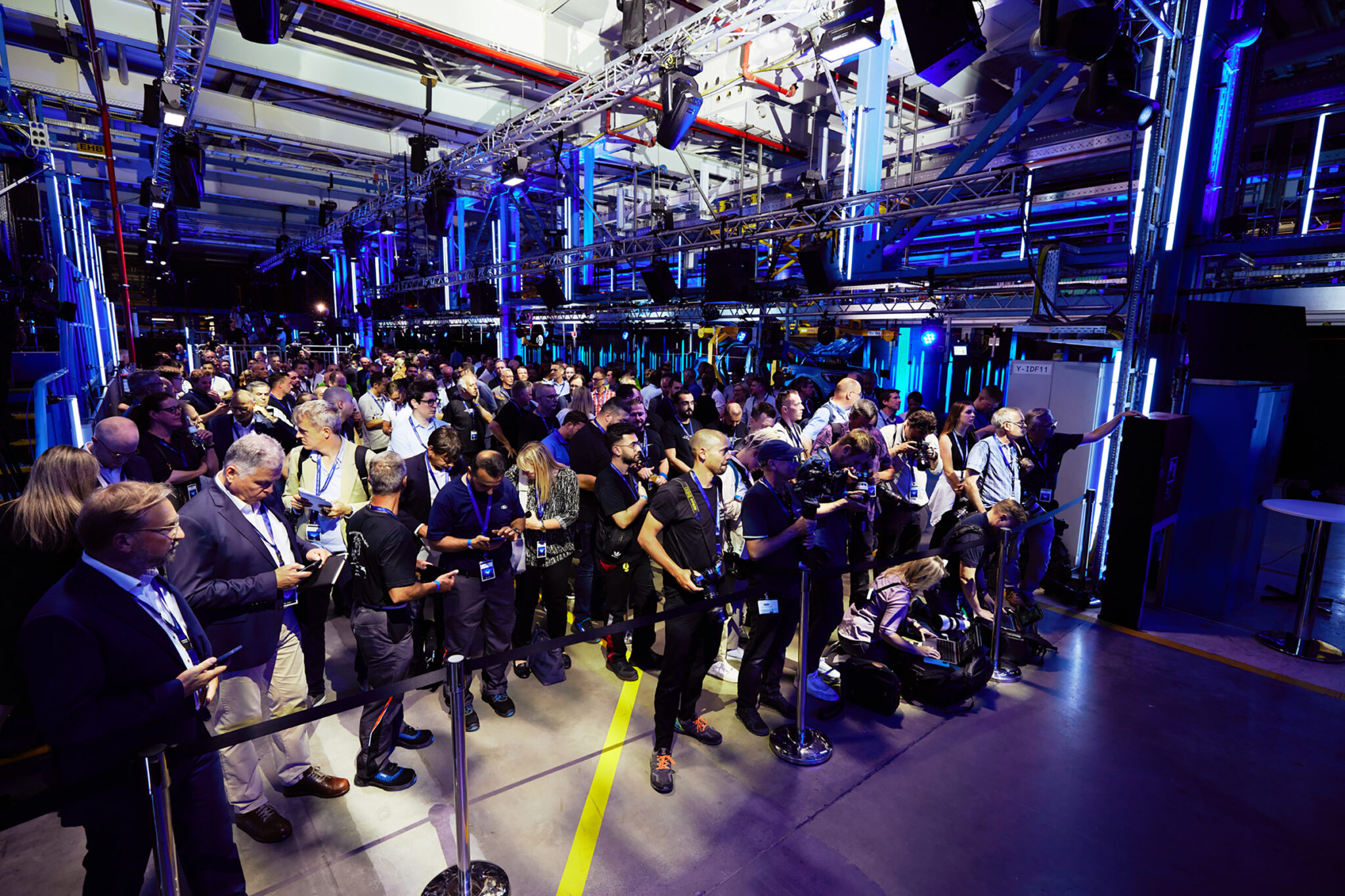 A dense crowd of seated and standing guests fills an industrial hall, lit in blue and violet with overhead gantries and pipework visible above.