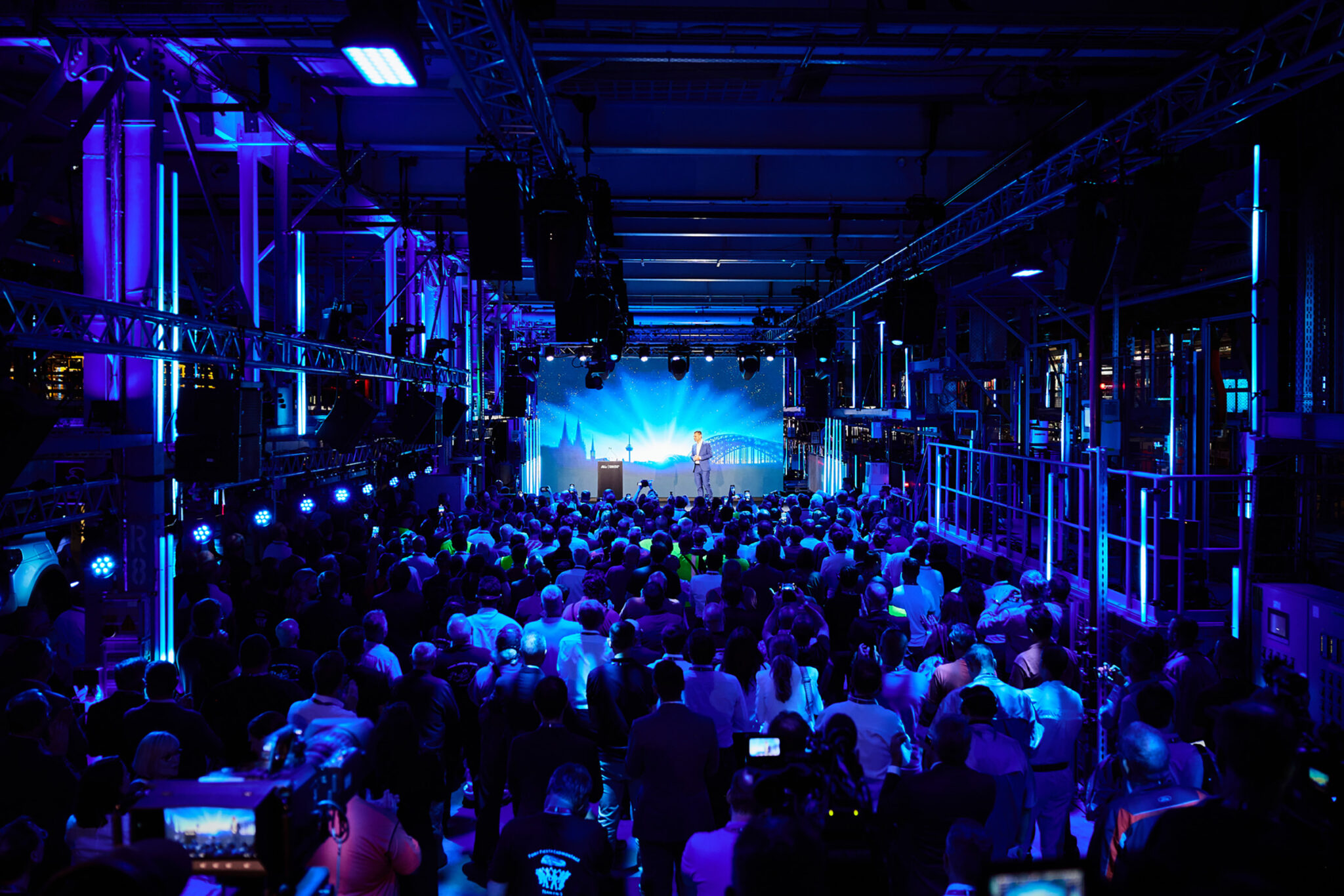 A large seated audience fills a former factory hall lit in deep blue, facing a bright stage screen in the distance beneath exposed industrial roof structure.