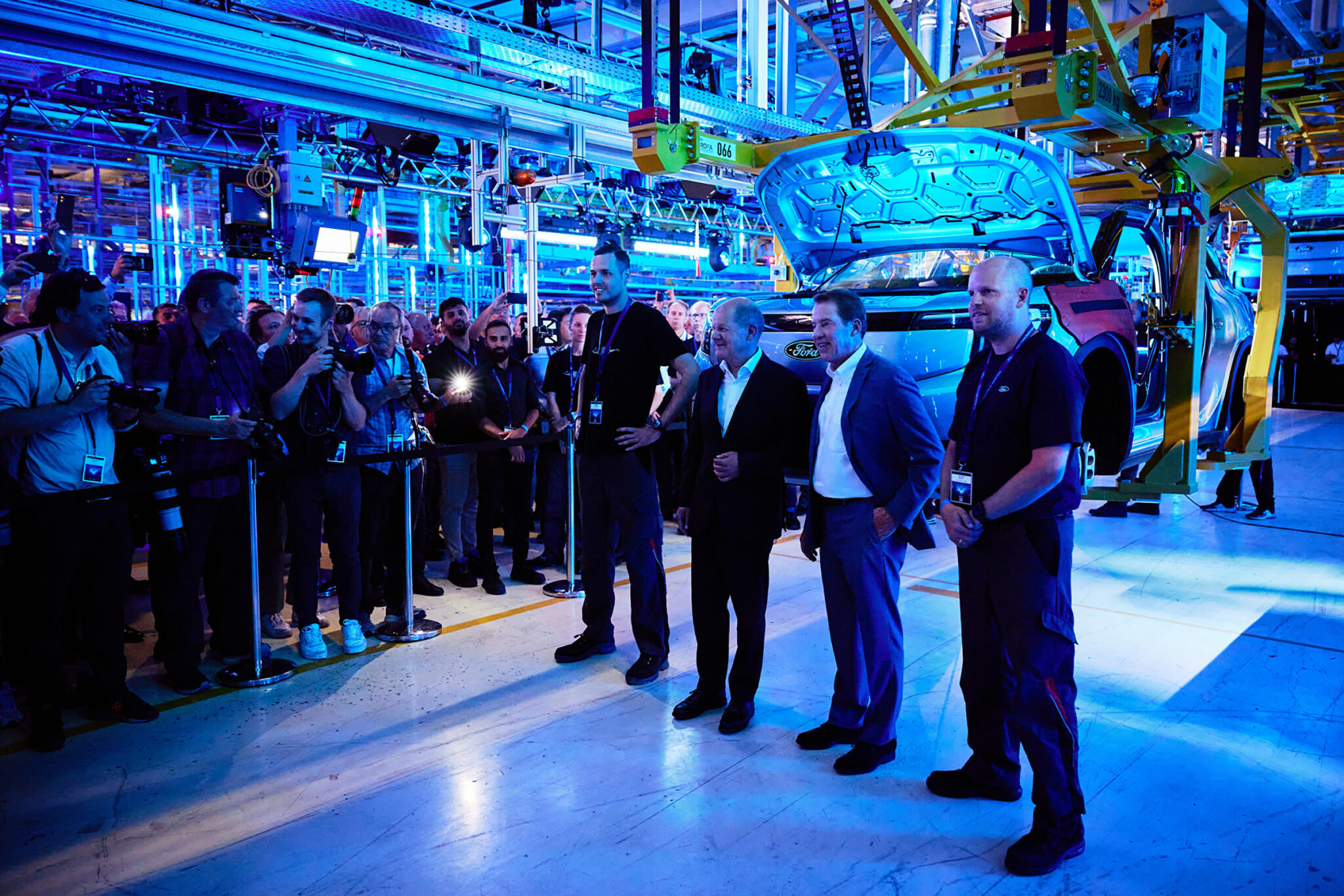 A group of people in suits stands talking on a glossy blue-lit floor inside a large industrial hall, with machinery and bright lighting behind them.