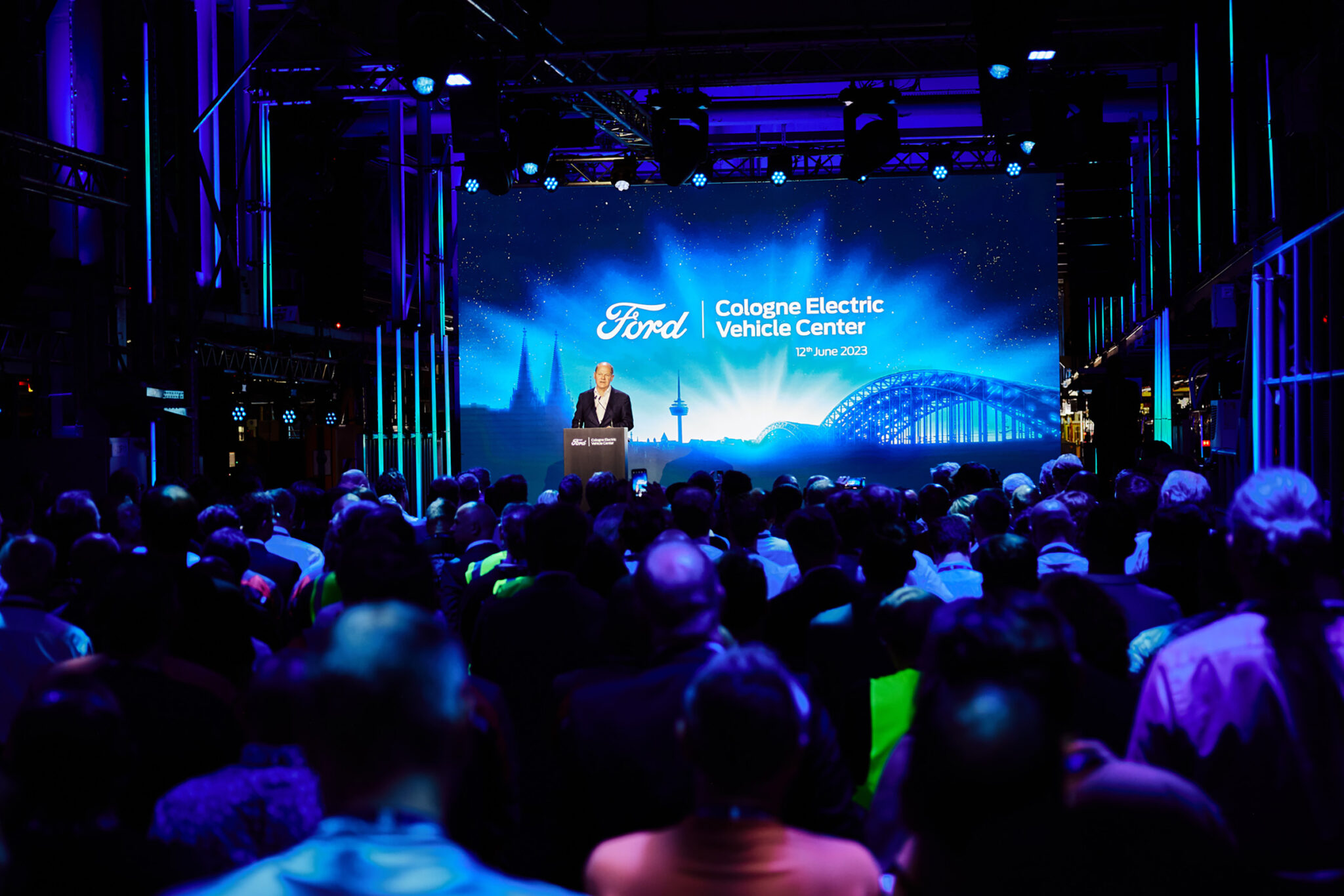 A wide stage with a large bright screen at the centre of a darkened industrial hall, with a seated audience in silhouette in the foreground.