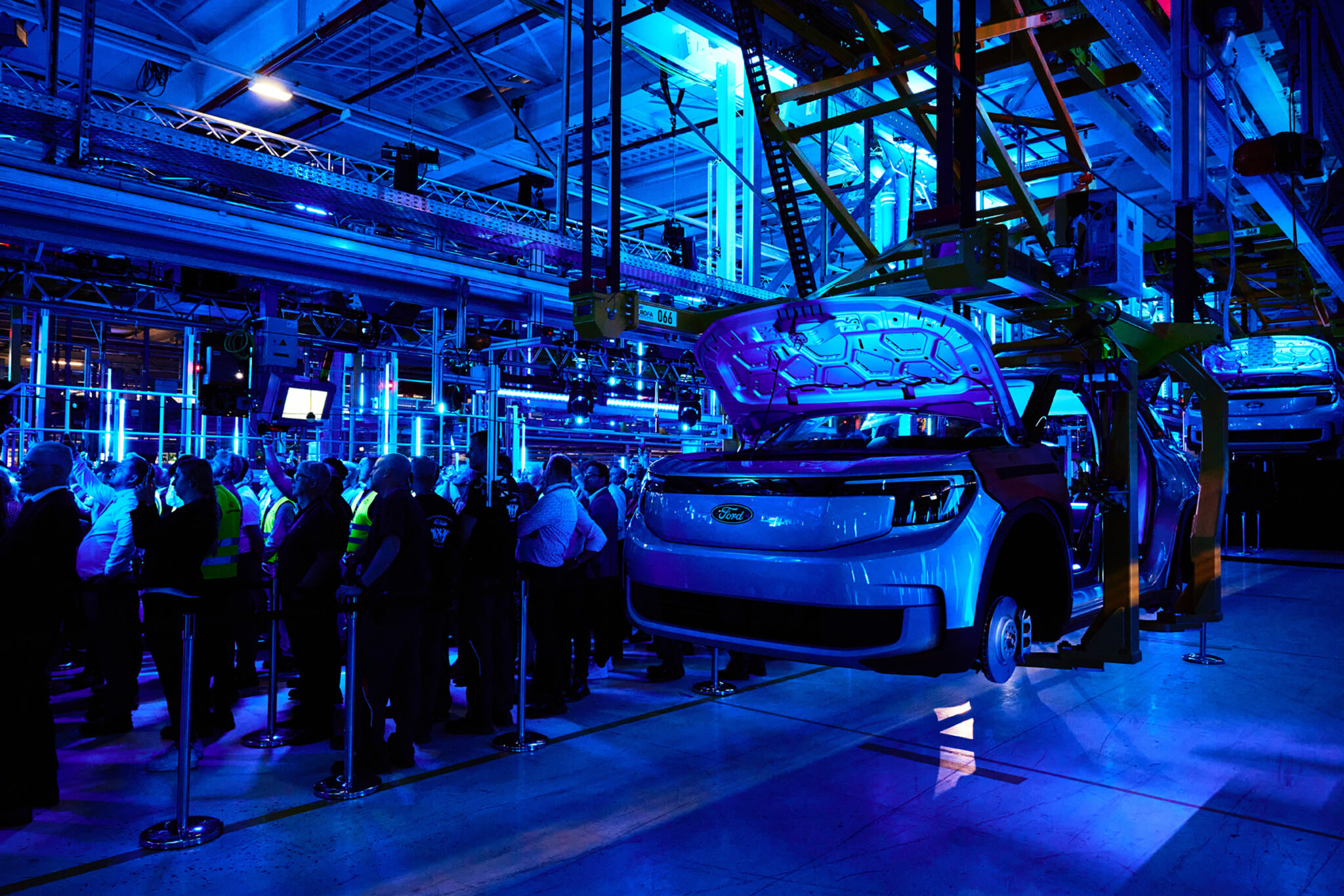 A vehicle stands on a factory production line surrounded by machinery and overhead rigging, with a crowd of visitors gathered along the edges in blue light.