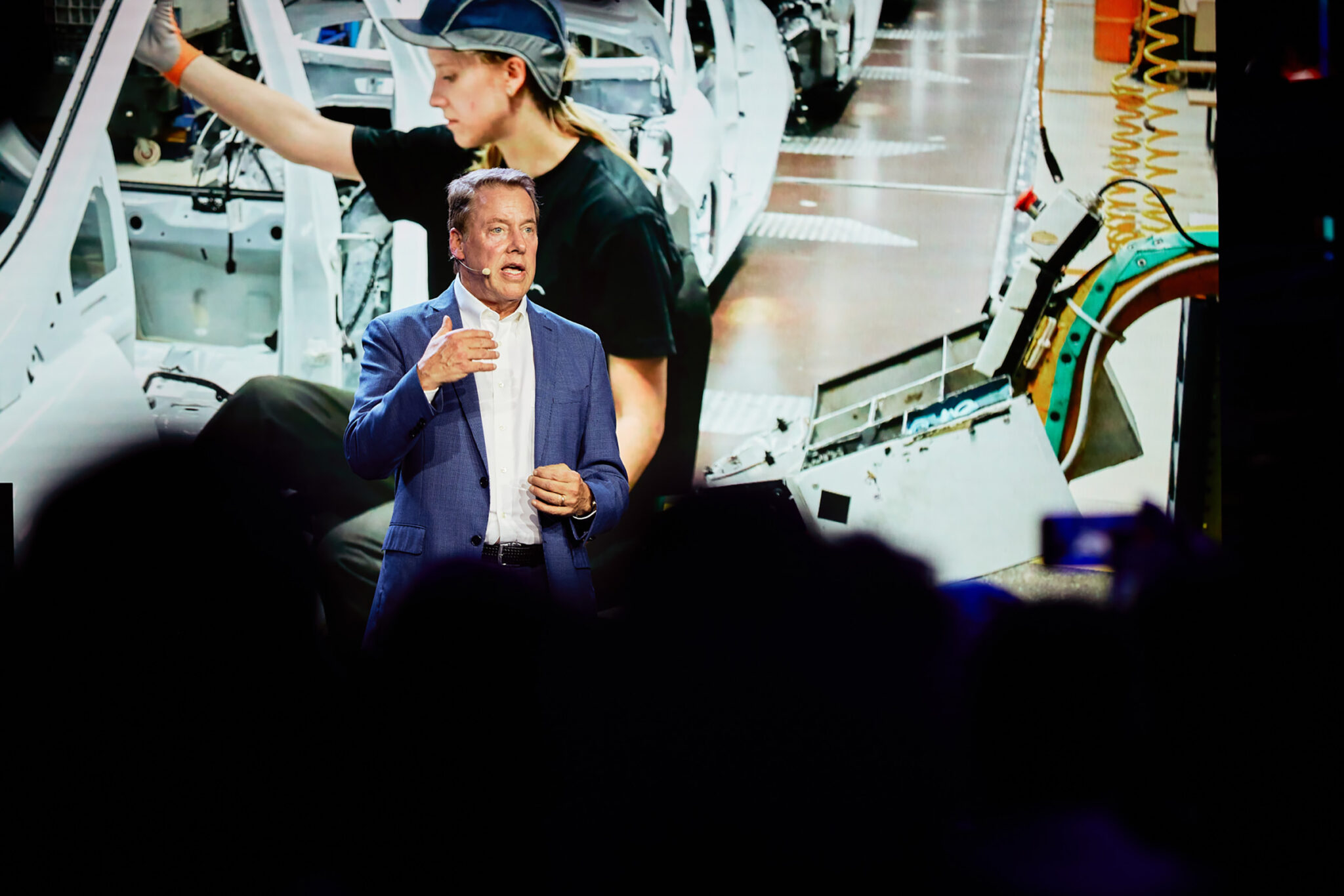 A man in a dark jacket speaks and gestures with one hand raised, standing in front of a brightly lit backdrop of vehicle body parts and machinery.