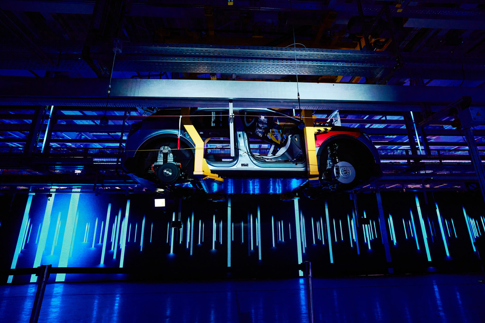 A bare electric vehicle chassis and battery platform stands on a plinth, lit from below in blue with yellow highlights picking out its components.
