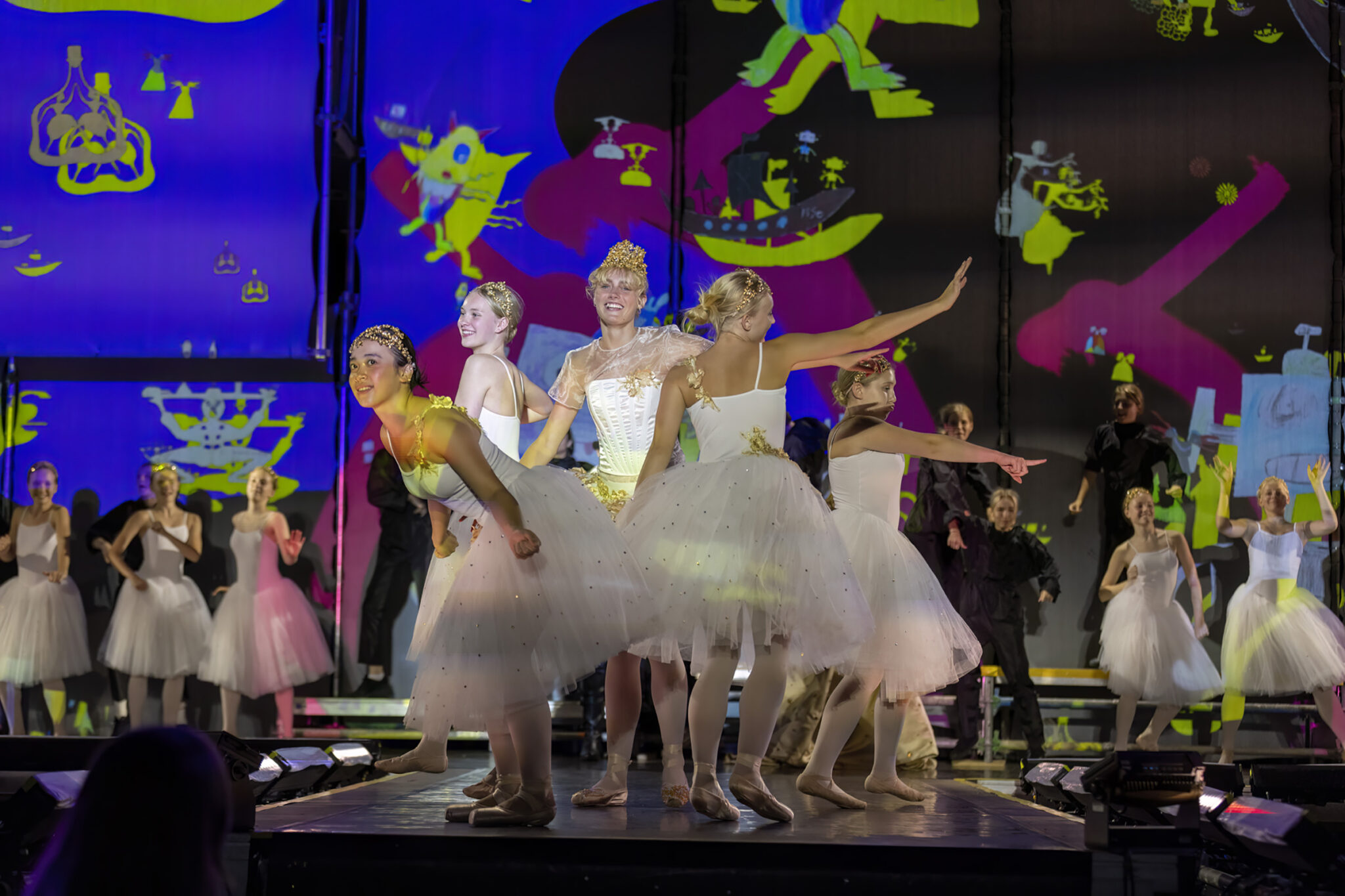 Dancers in white tutus stand in a row on a stage lit in blue and green, with a bright projected pattern behind them.