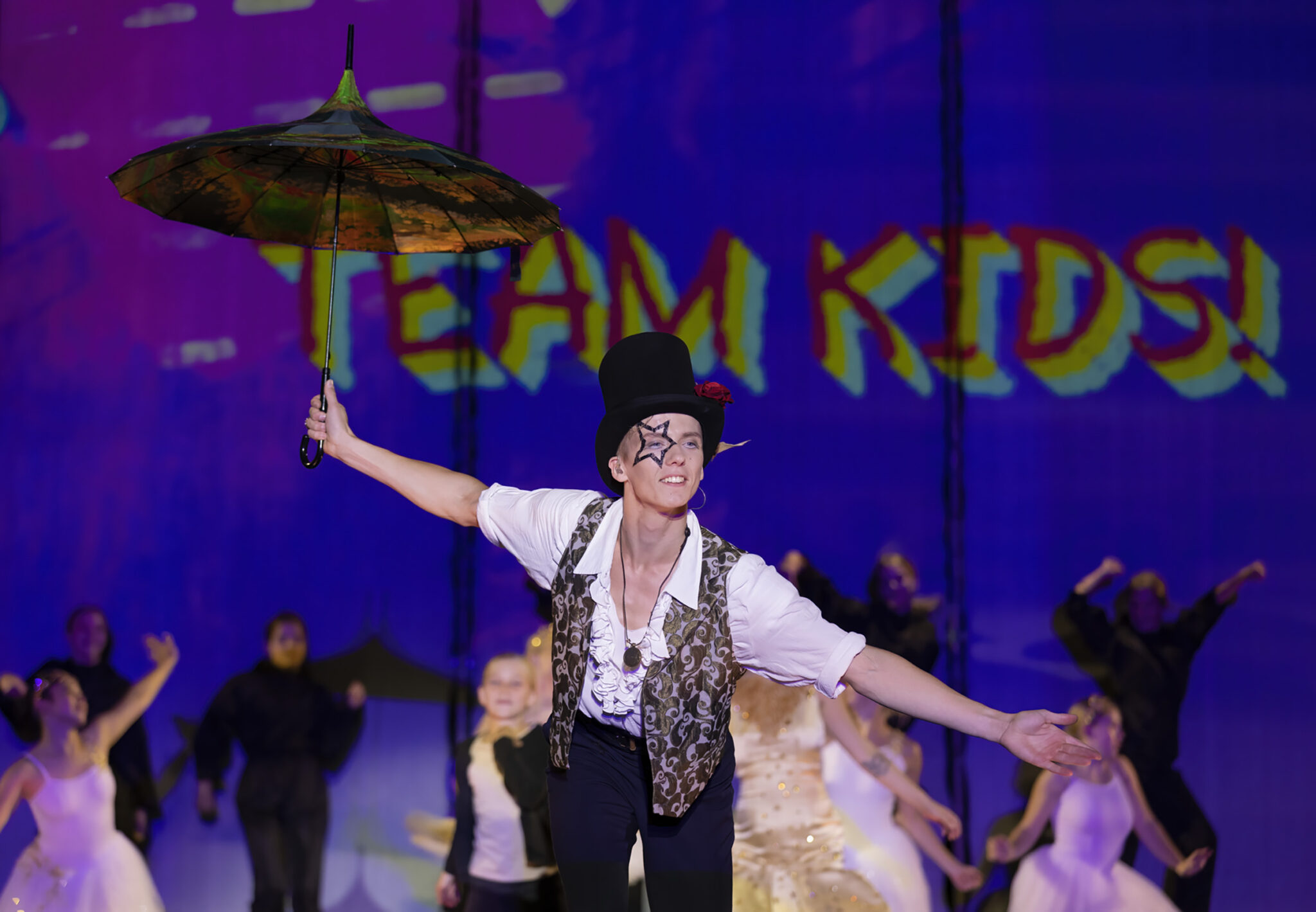 A performer in a dark costume and hat leans back mid-movement holding a parasol, with other dancers behind on a stage lit in purple and blue.