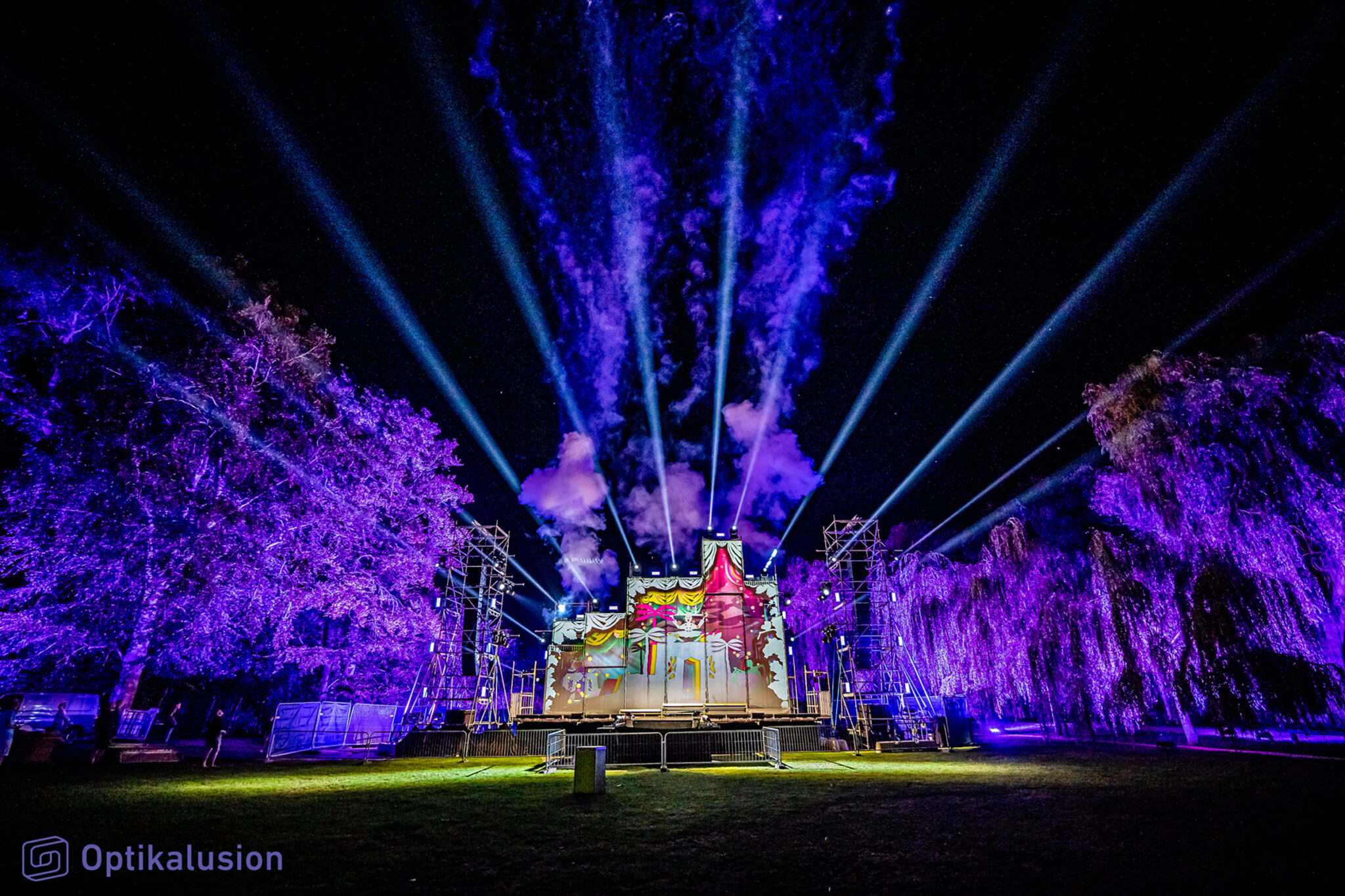 An outdoor stage at night lit in deep violet and blue, with a decorated set at its centre and tall trees on either side picked out by coloured light.
