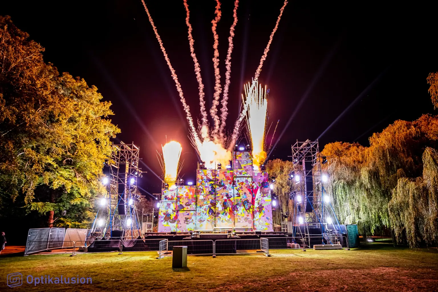 Fireworks rise in white streaks above a brightly lit outdoor stage at night, with autumn trees on either side and an audience area in front.
