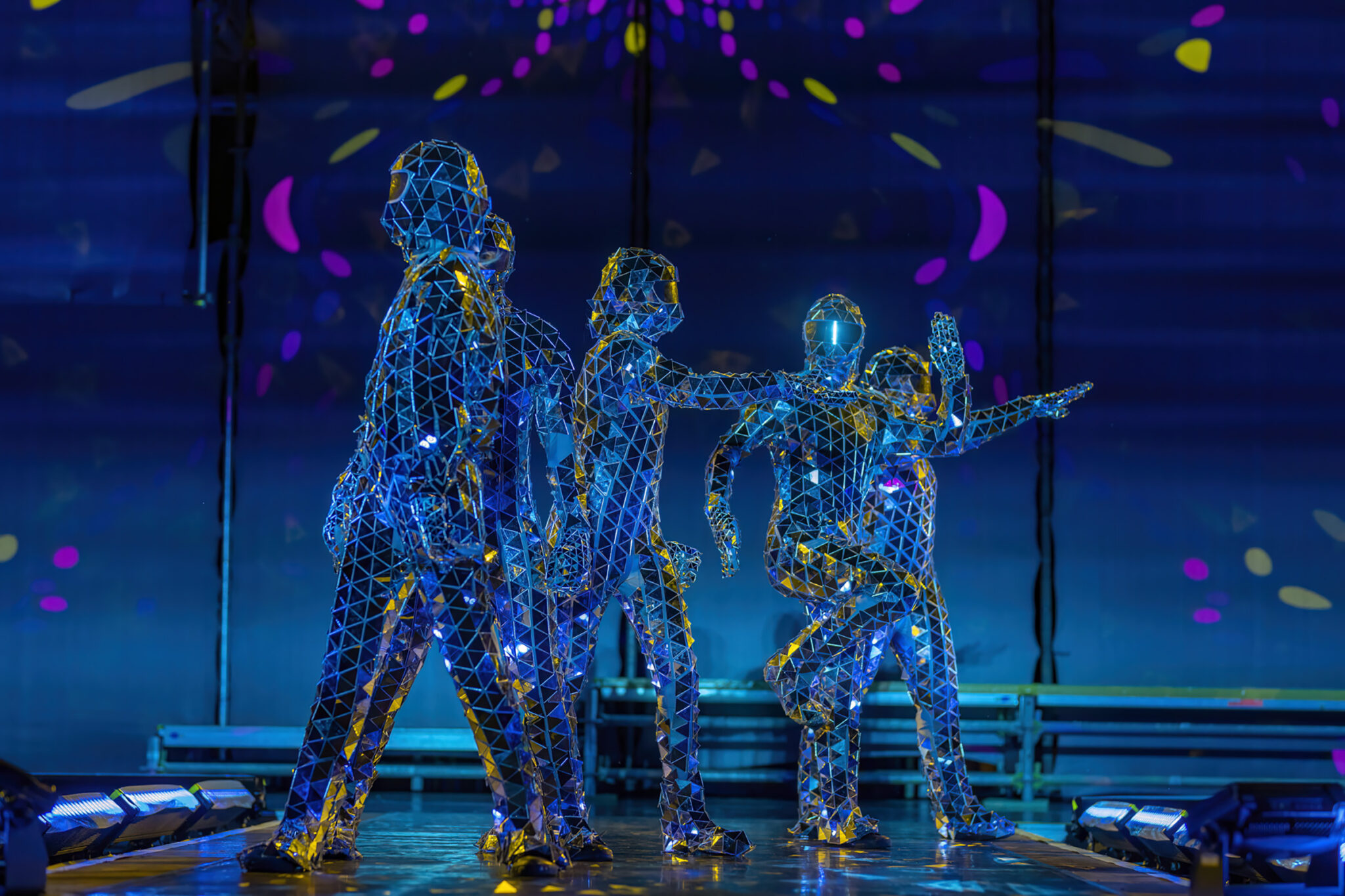 Two performers in illuminated wire-frame costumes stand on a darkened stage, their outlines traced in bright blue light against a deep blue background.