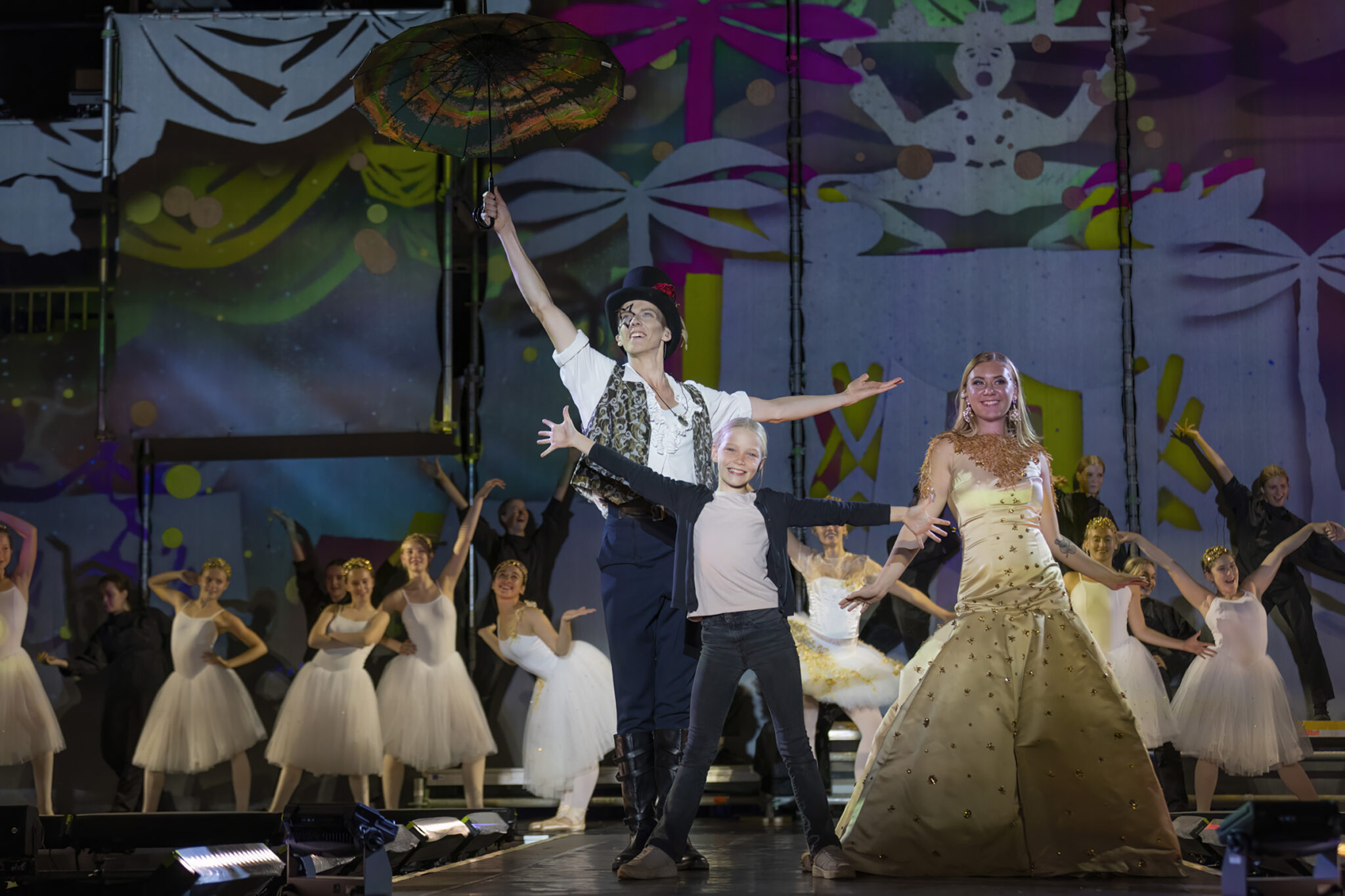 Two performers stand centre stage with arms raised, one in a long pale gown, surrounded by a group of dancers in white against a dark painted backdrop.