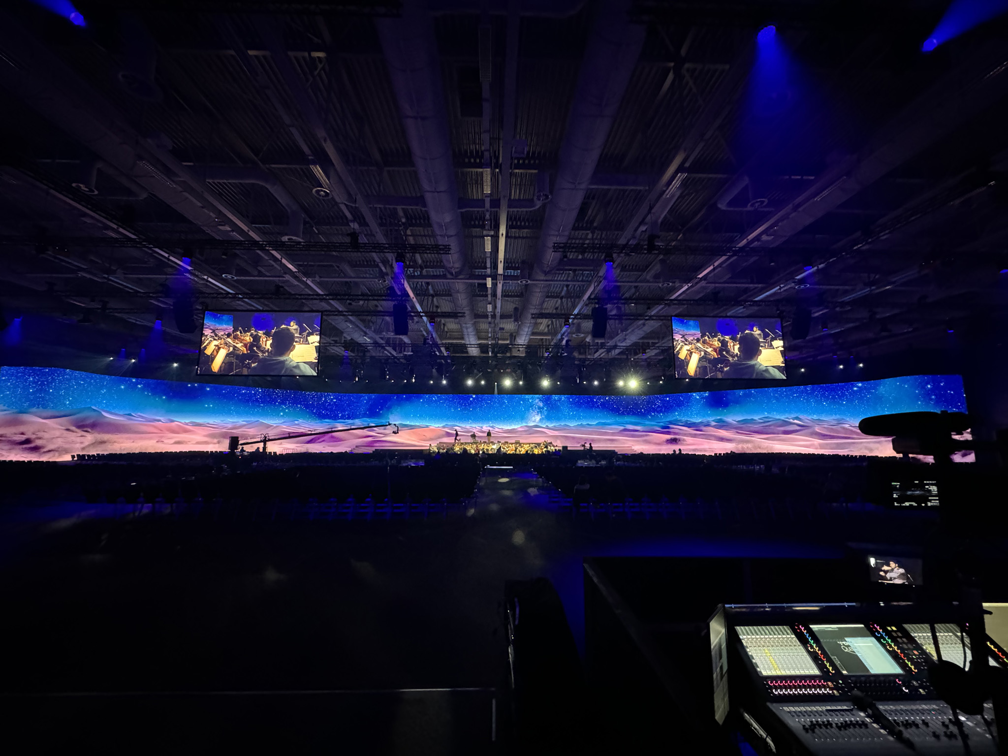 A panoramic LED wall shows a starry night sky over desert dunes, with an orchestra on stage, two hanging screens showing musicians and a lit mixing desk in the foreground.