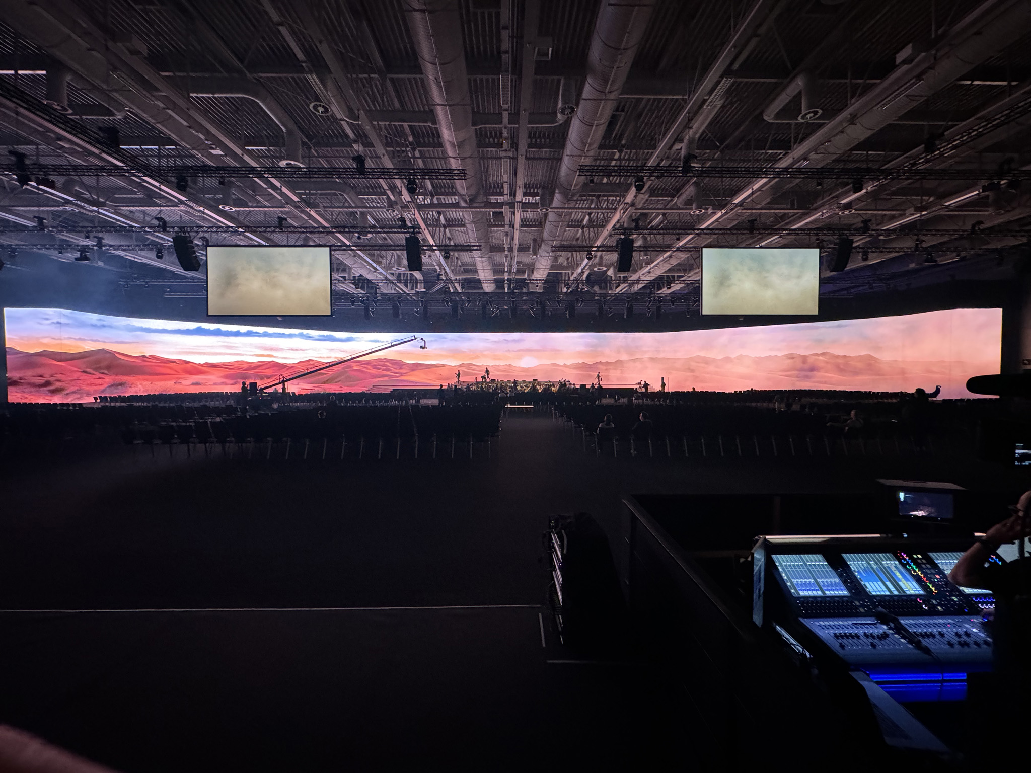 A huge curved LED wall shows a pink desert at sunset across a dark exhibition hall, with rows of empty seats, hanging screens and a mixing desk in the foreground.