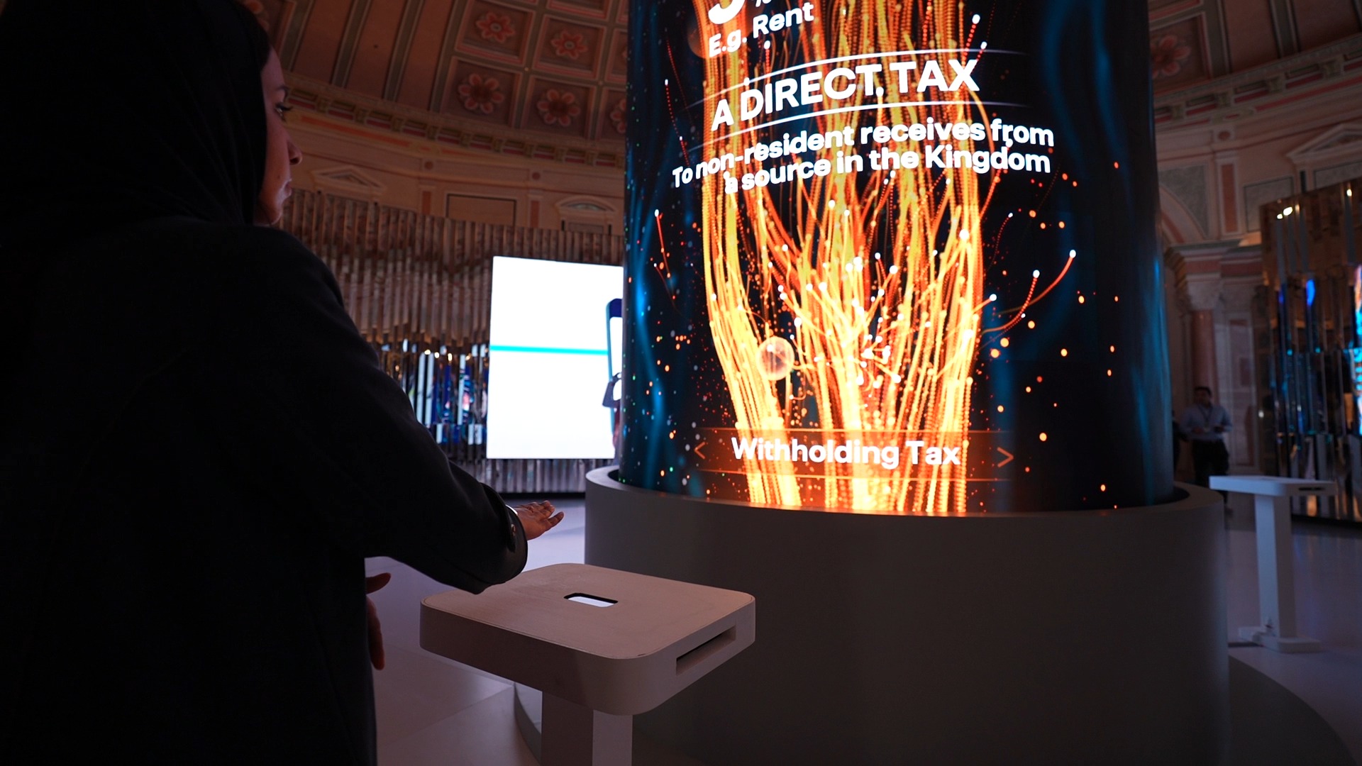 A visitor stands at a small white kiosk beside a cylindrical LED tower showing orange streams of light and English text.