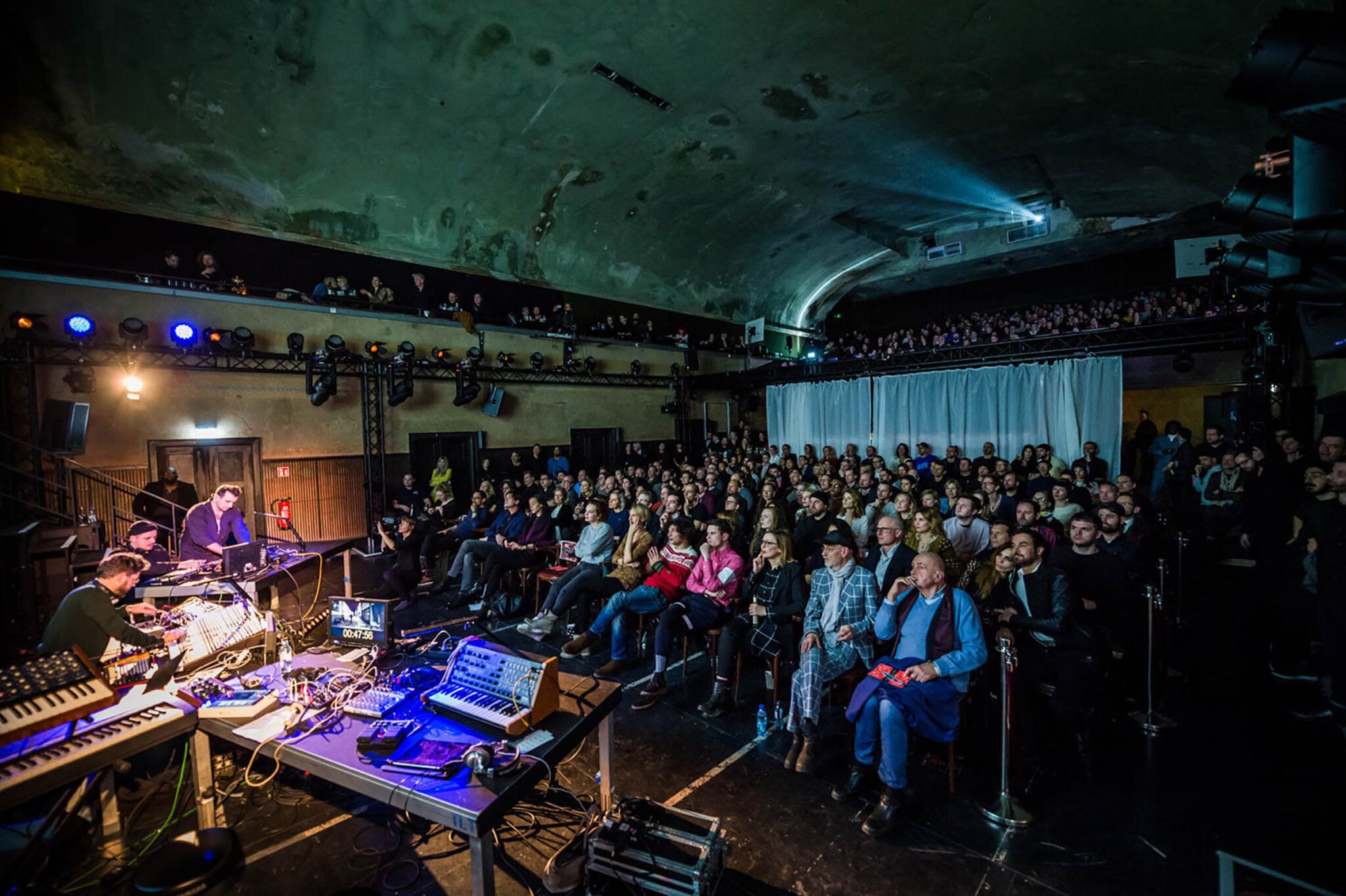 A full cinema auditorium photographed from the stage, the audience seated in tiered rows behind a foreground table of synthesisers, laptops and mixing equipment.