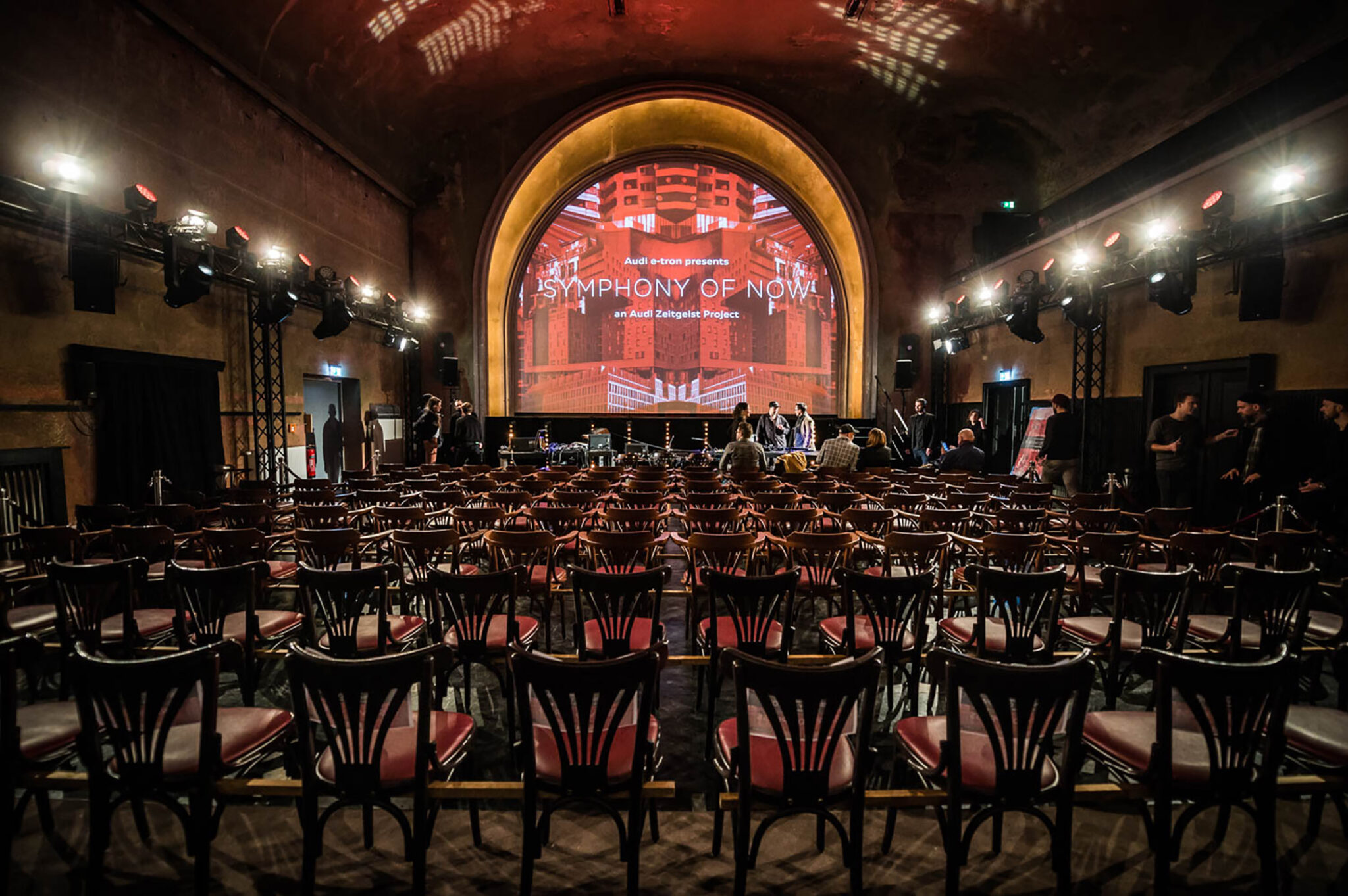 Empty cinema auditorium with curved rows of dark bentwood chairs facing a large arched screen that shows an illustrated red city skyline, framed by rows of stage lights.