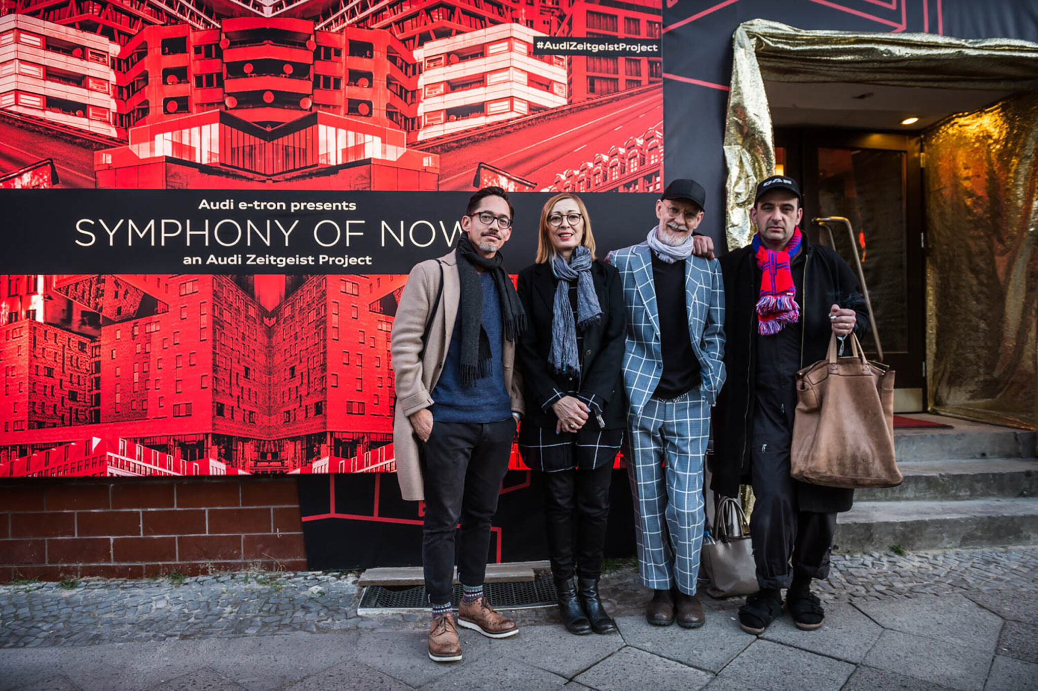Four people in winter coats standing together for a photograph in front of a large red backdrop printed with an illustrated city skyline.