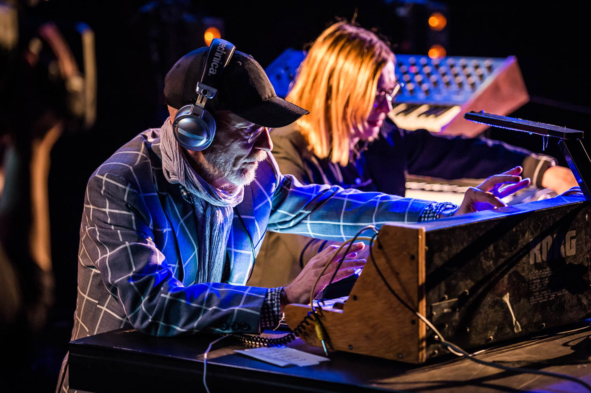 Two musicians playing synthesisers side by side on a dark stage, blue projected light patterning their faces, hands and instruments.