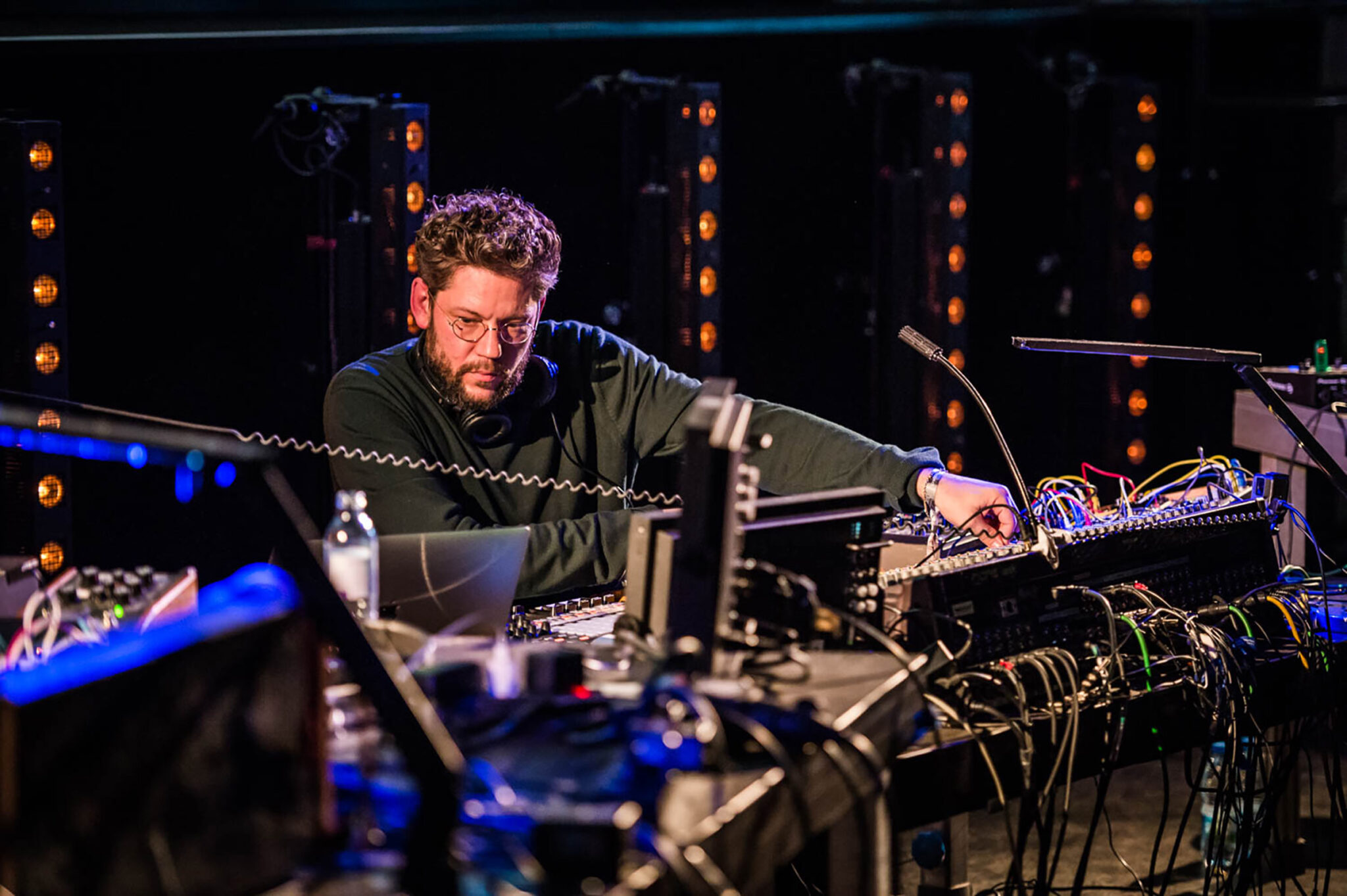 A musician working at a modular synthesiser rig densely strung with patch cables, lit from behind by vertical rows of warm filament bulbs.