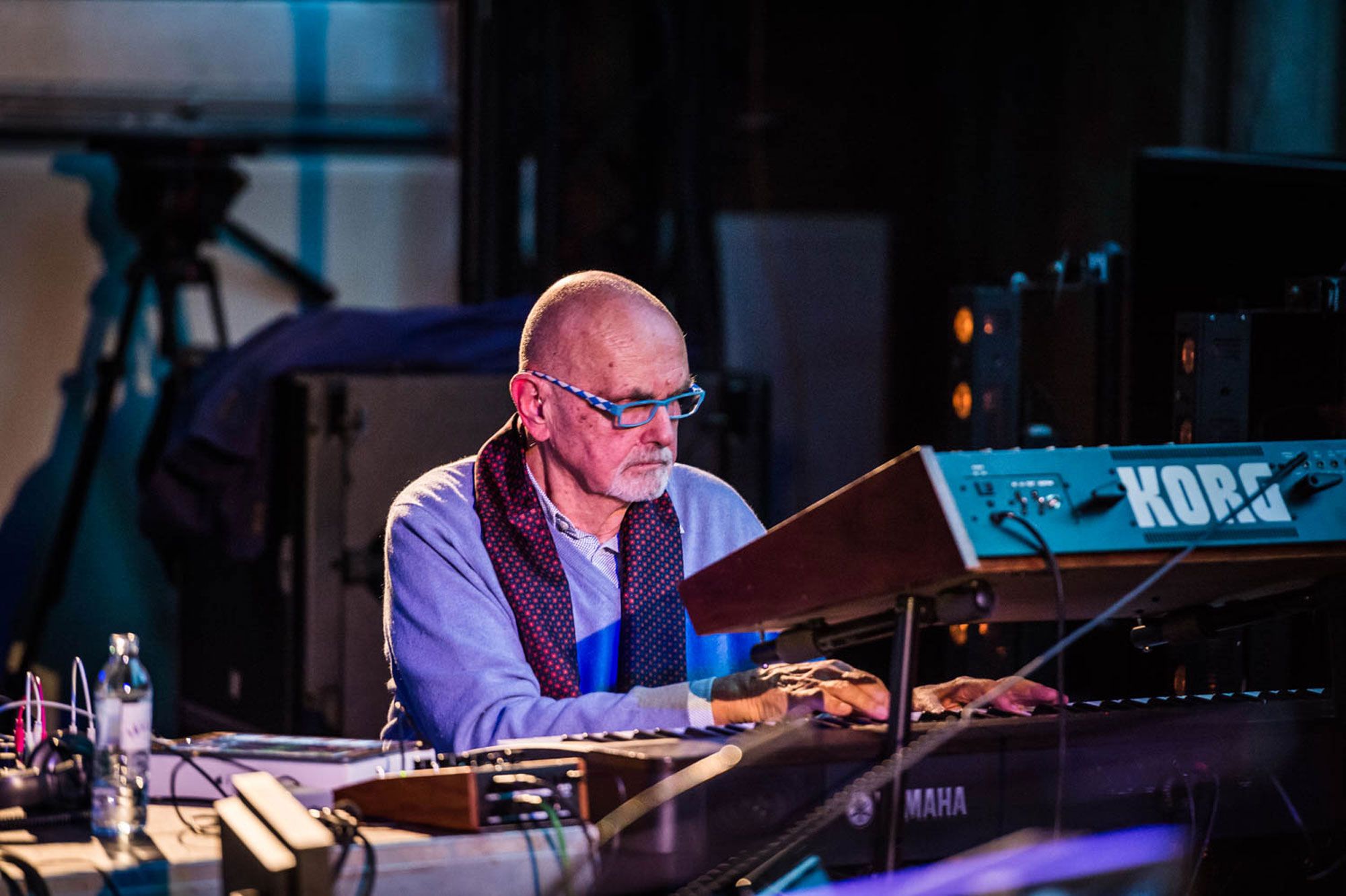 A musician seated at a keyboard synthesiser on a darkened stage, reading from a lit music stand while playing.