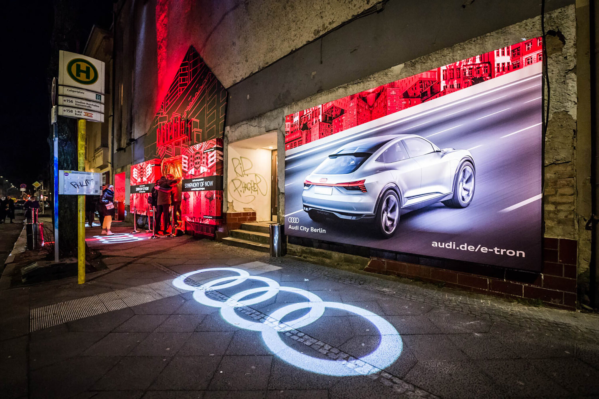A night street beneath a railway bridge, with a large illuminated billboard showing a car and four interlocking rings projected in light onto the wet pavement.