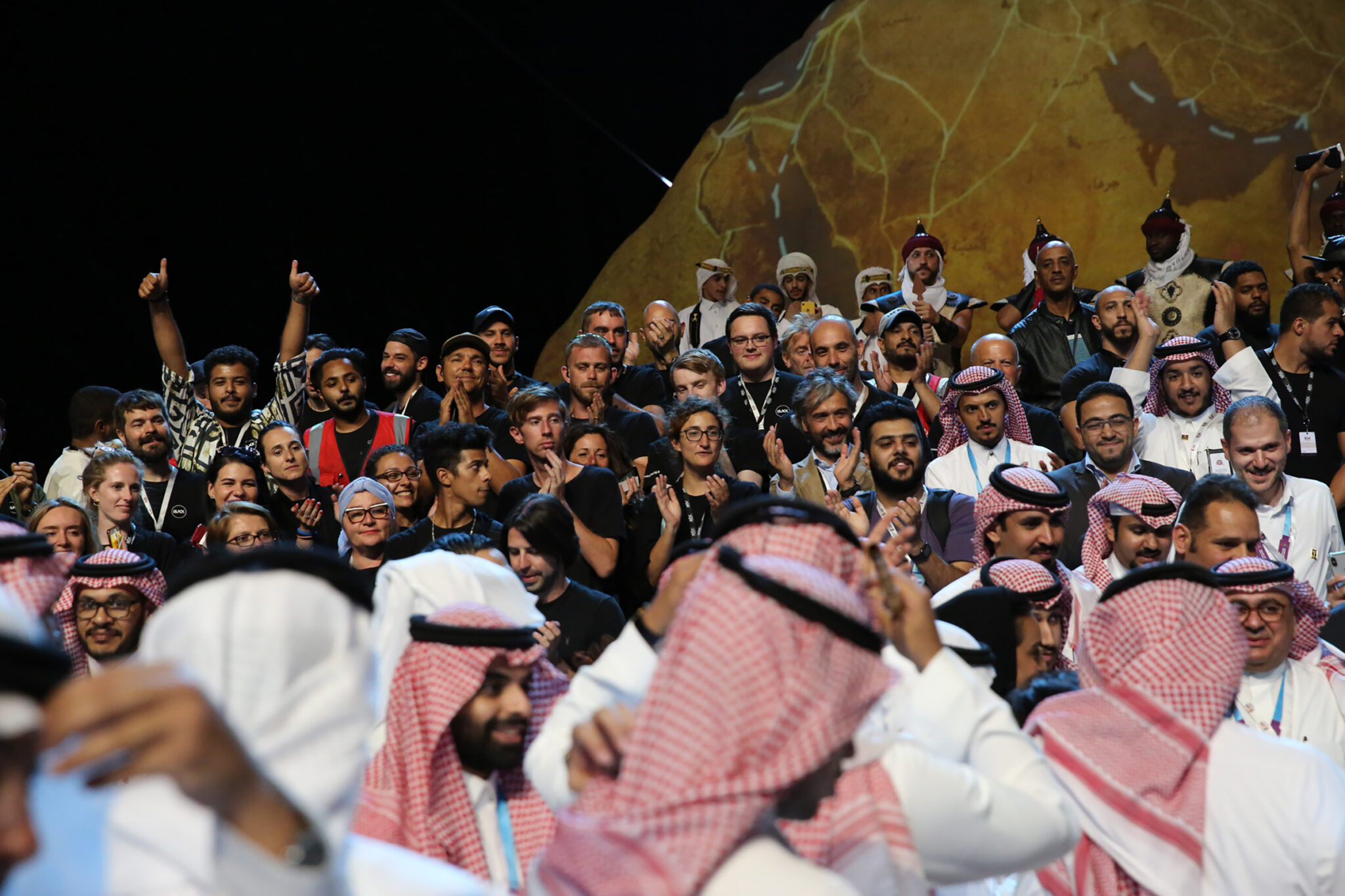 A packed audience in white robes and headdresses, many with arms raised and phones held up, lit in warm light.